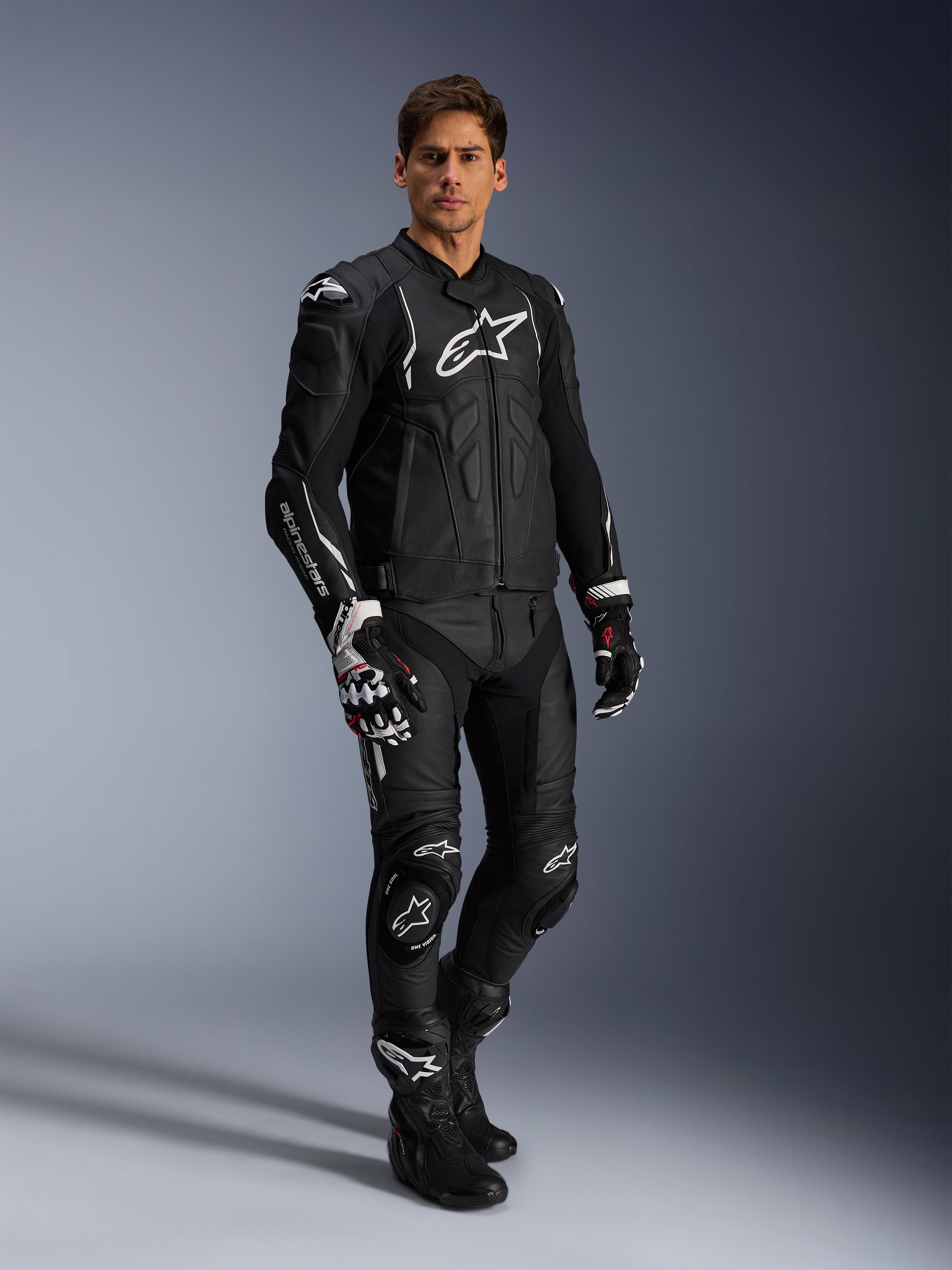 Męski motocyklista ubrany w skórzaną kurtkę Alpinestars Dusk w kolorze czarno-białym, kurtka premium z dużym białym logo na klatce piersiowej, w zestawie z dopasowanymi skórzanymi spodniami wyścigowymi, rękawicami i butami, kompletny sportowy strój motocyklowy