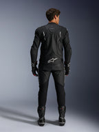 Motocyklista widziany od tyłu w kurtce skórzanej Alpinestars Dusk i spodniach Missile V3, czarny skórzany zestaw z białymi akcentami, kurtka z napisem „Dusk” i logo Alpinestars na plecach, czarne buty i rękawice wyścigowe, profesjonalny strój motocyklowy do jazdy szosowej.