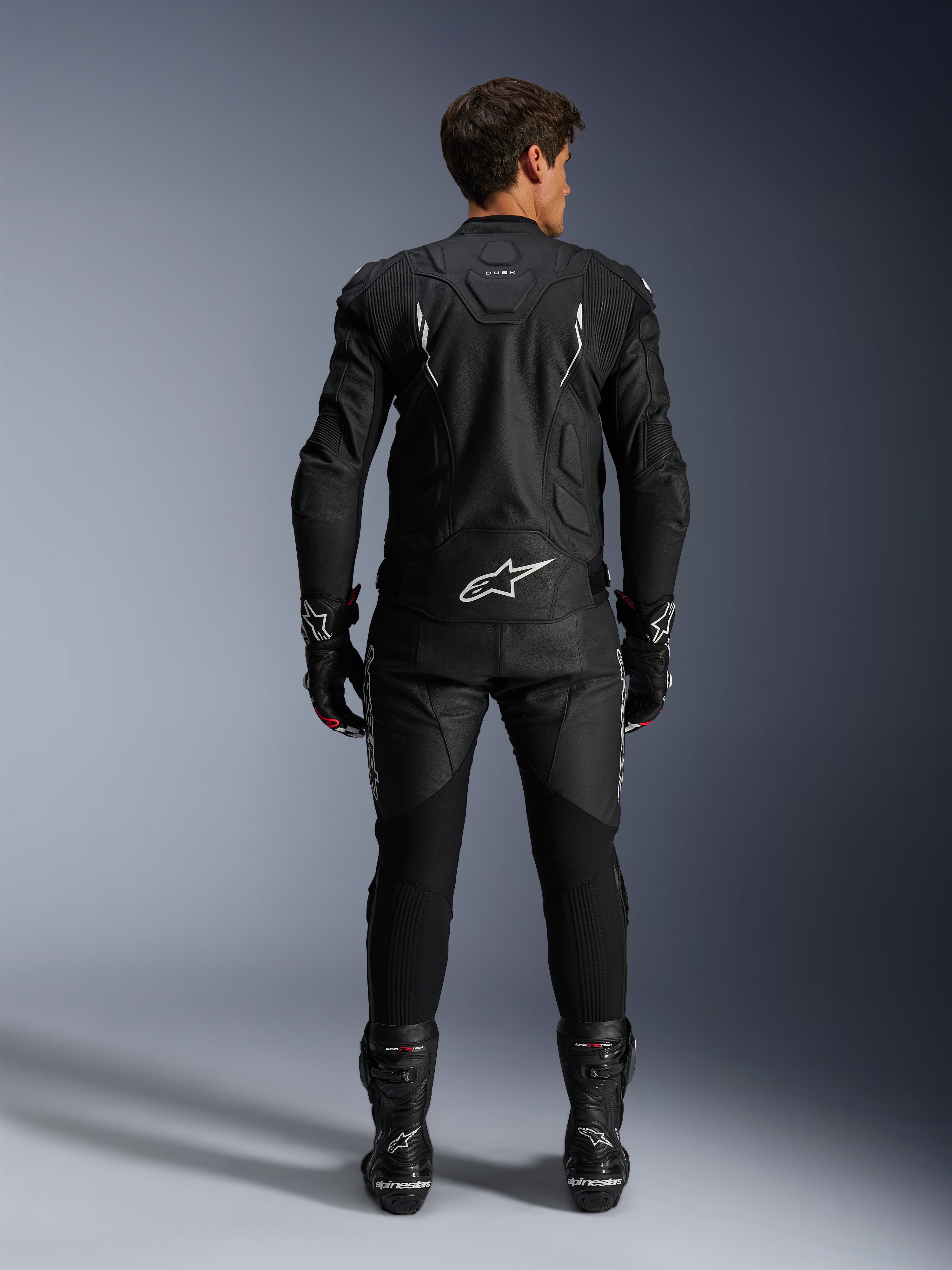 Motocyklista widziany od tyłu w kurtce skórzanej Alpinestars Dusk i spodniach Missile V3, czarny skórzany zestaw z białymi akcentami, kurtka z napisem „Dusk” i logo Alpinestars na plecach, czarne buty i rękawice wyścigowe, profesjonalny strój motocyklowy do jazdy szosowej.