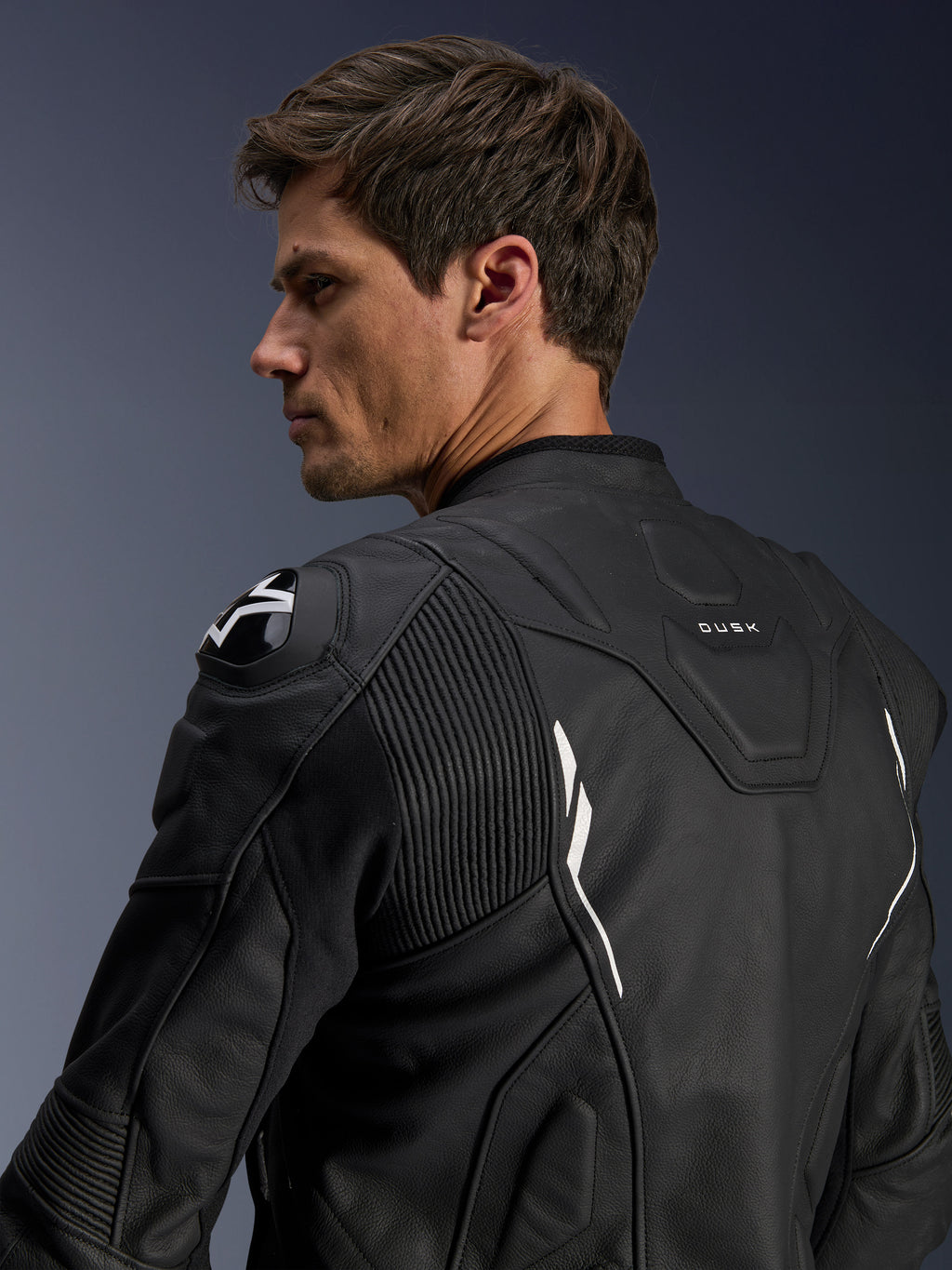 Skórzana kurtka Alpinestars Dusk, czarna z białymi akcentami, widok z tyłu na modelu, ochraniacze ramion DFS z logo Alpinestars, skórzane panele harmonijkowe zwiększające swobodę ruchów, branding „DUSK” na górze pleców, kurtka motocyklowa z najwyższej jakości skóry bydlęcej do jazdy szosowej i sportowej