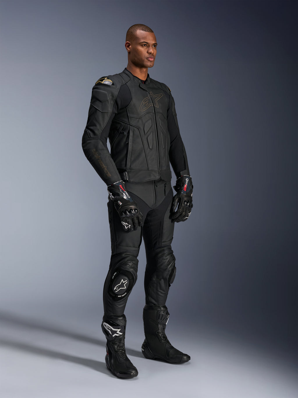 Męski model w skórzanej kurtce Alpinestars Dusk w kolorze Black Laser, zestawionej z pasującymi czarnymi skórzanymi spodniami wyścigowymi, rękawicami GP Pro i wyczynowymi butami torowymi. Pełny motocyklowy strój wyścigowy z zewnętrznymi ochraniaczami ramion DFS i sportowym dopasowaniem do jazdy szosowej i sportowej.