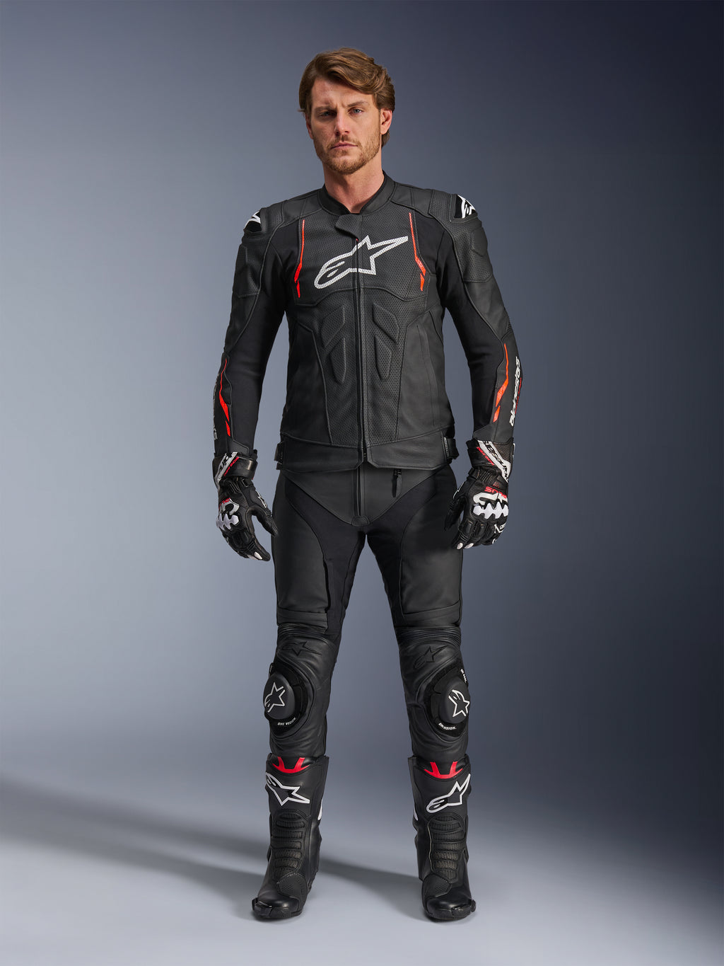 Motocyklista w skórzanej kurtce Alpinestars Dusk Airflow w kolorze Black Red Fluo, z perforowanymi panelami i białym logo na piersi, w zestawie z czarnymi skórzanymi spodniami, sliderami na kolanach, rękawicami i butami wyścigowymi, kompletny strój motocyklowy