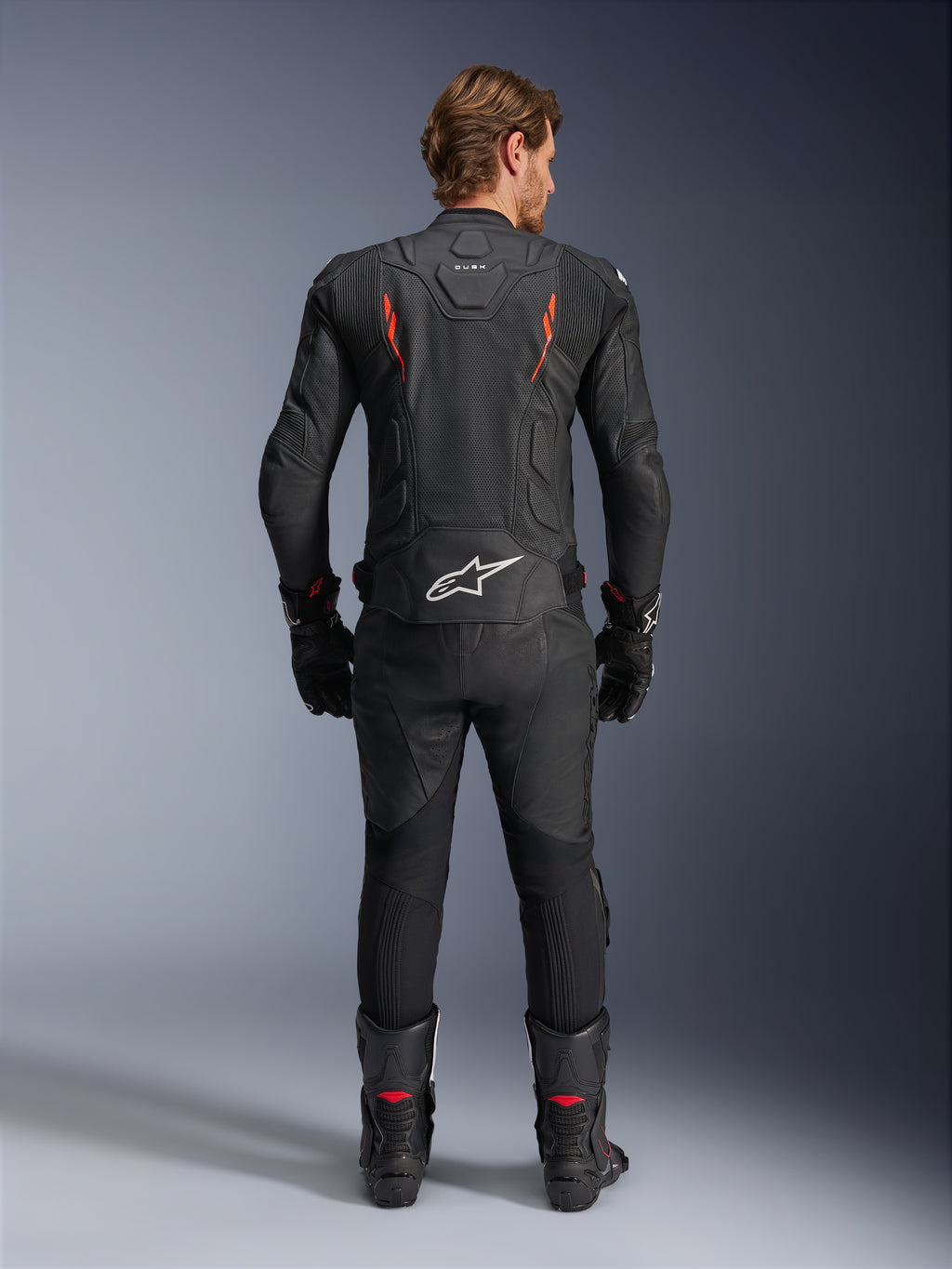 Skórzana kurtka Alpinestars Dusk Airflow, czarna i czerwona fluo, widok z tyłu ukazujący rozległe perforacje i duże białe logo w dolnej części pleców, noszona z pasującym skórzanym strojem wyścigowym, w tym spodniami, butami i rękawicami