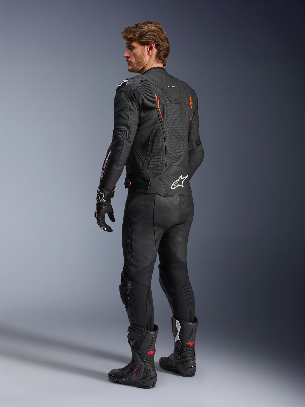 Skórzana kurtka Alpinestars Dusk Airflow, czarna z akcentami Red Fluo, widok z tyłu motocyklisty w pełnym skórzanym stroju motocyklowym, czarne spodnie, rękawice i buty wyścigowe, perforowana skóra, białe logo Alpinestars