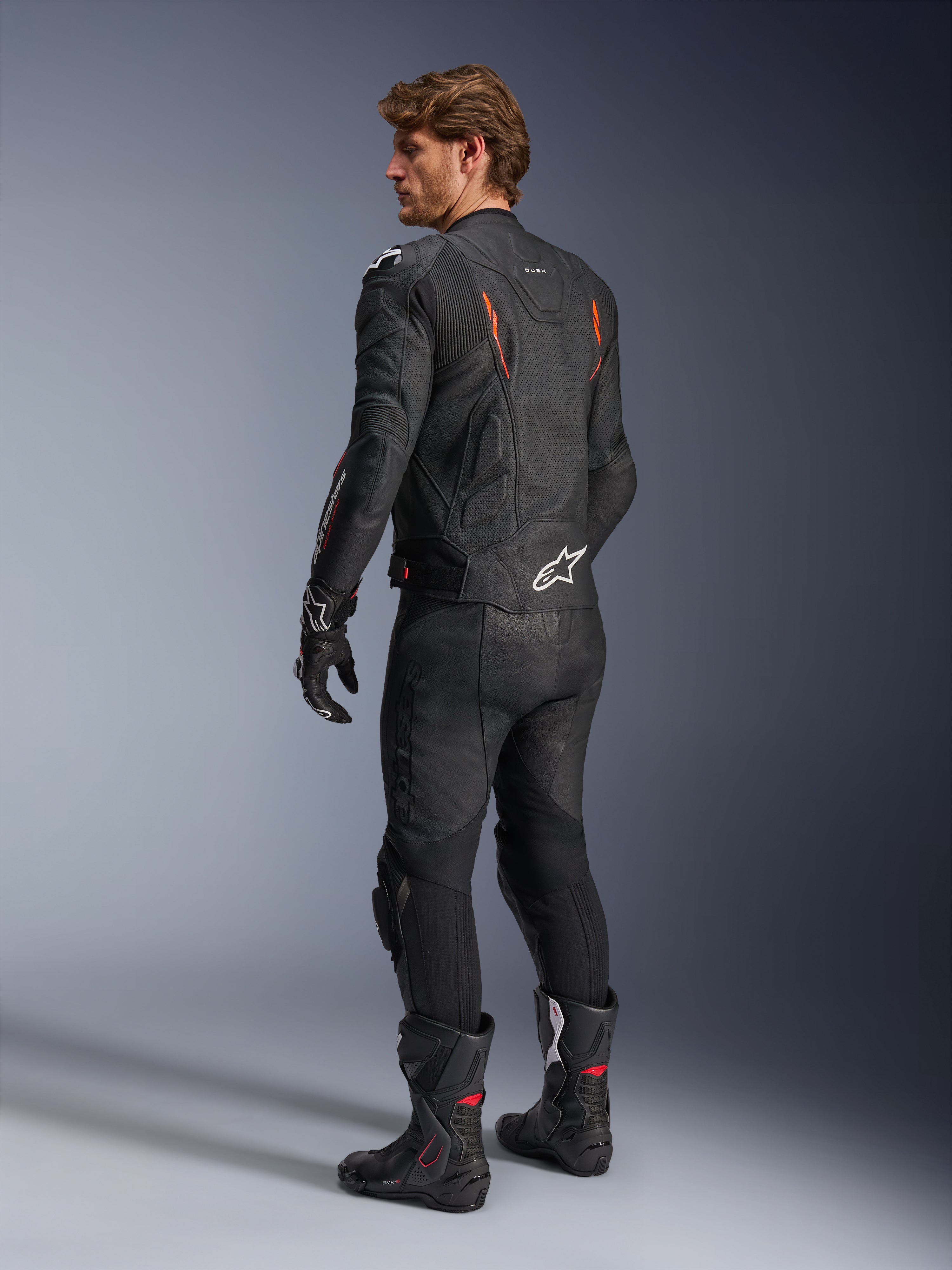 Skórzana kurtka Alpinestars Dusk Airflow, czarna z akcentami Red Fluo, widok z tyłu motocyklisty w pełnym skórzanym stroju motocyklowym, czarne spodnie, rękawice i buty wyścigowe, perforowana skóra, białe logo Alpinestars