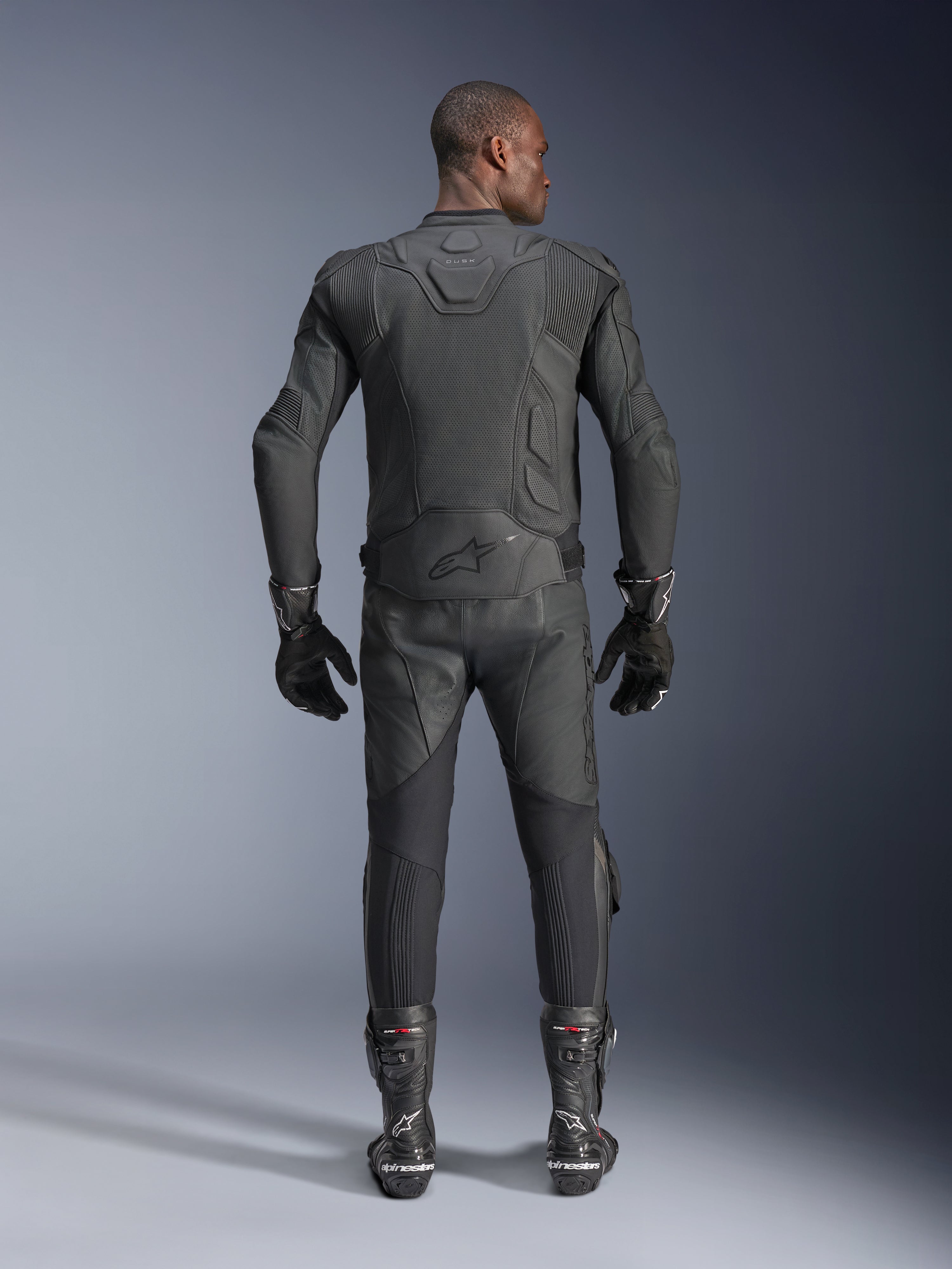 Męski model widoczny od tyłu w skórzanej kurtce Alpinestars Dusk Airflow w kolorze czarnym, w zestawie z dopasowanymi czarnymi skórzanymi spodniami wyścigowymi, rękawicami i butami, z licznymi perforacjami zapewniającymi wentylację oraz logo Alpinestars w dolnej części pleców.