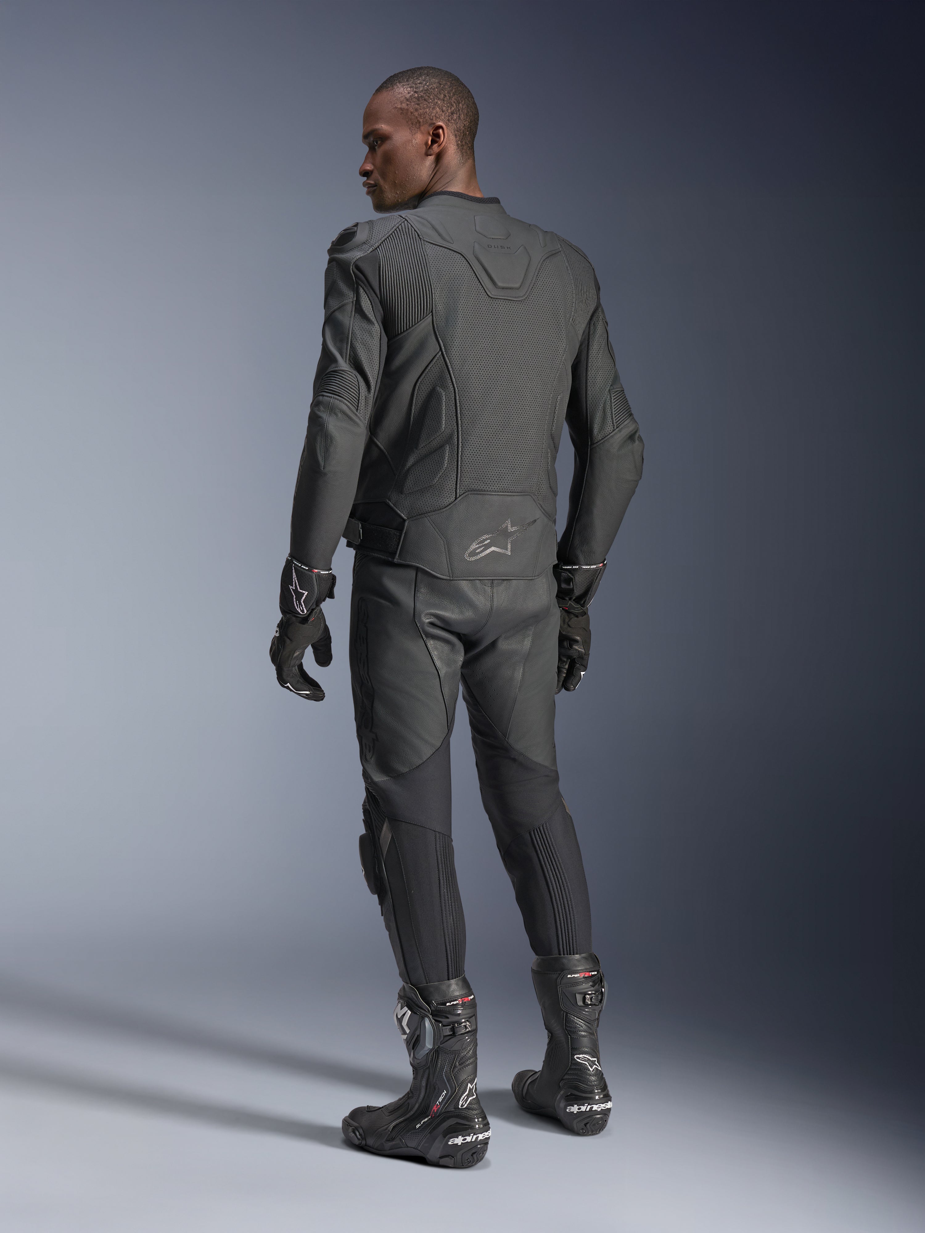Alpinestars Dusk Airflow Leather Jacket, motocyklista widziany od tyłu w czarnym skórzanym zestawie, widoczne rozległe perforacje na plecach, pasujące skórzane spodnie, rękawice i buty, sportowy krój