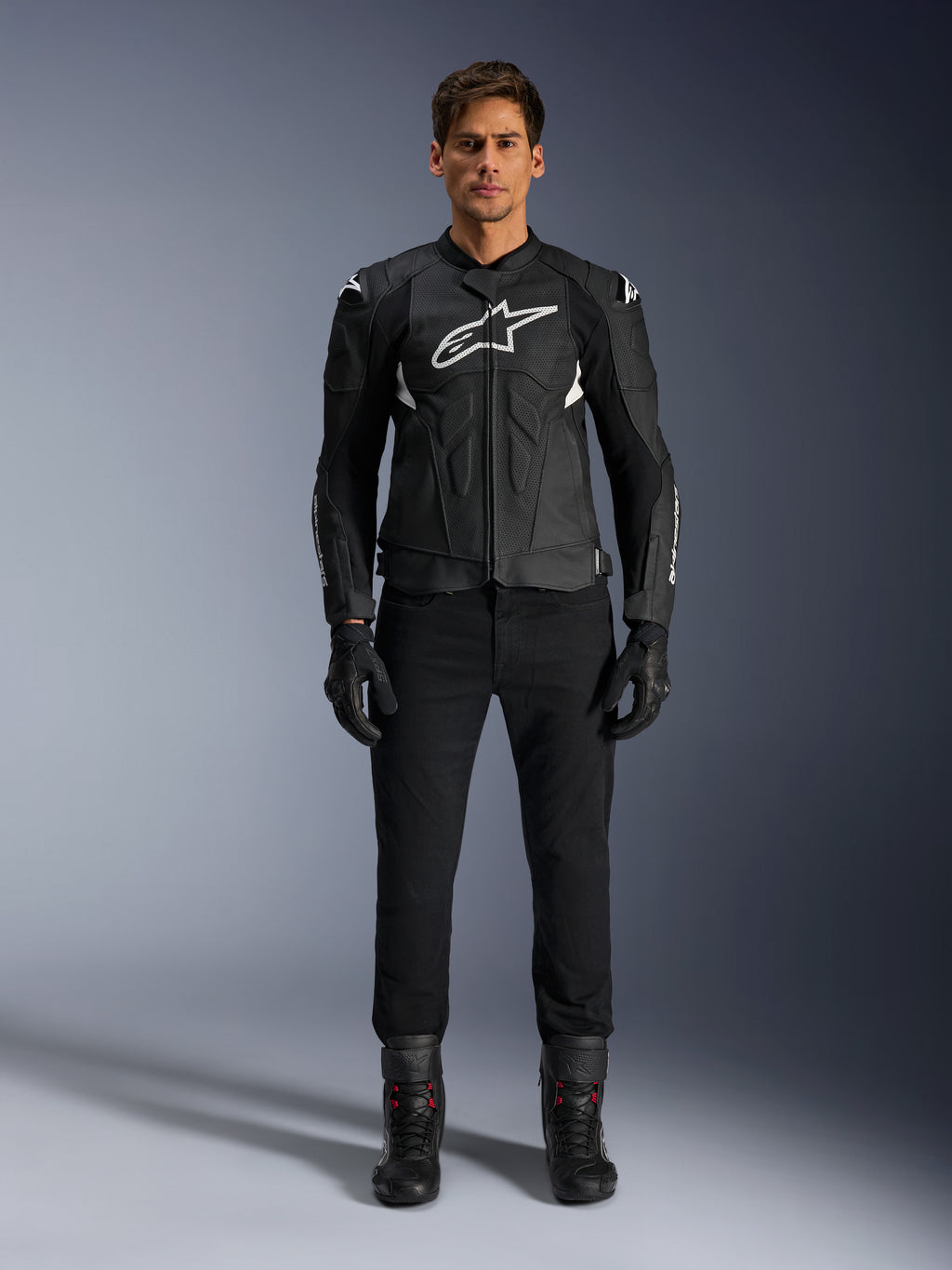 Alpinestars Dusk Airflow Leather Jacket, czarno-biała, motocyklowa kurtka skórzana z perforowanymi panelami dla przepływu powietrza, białe logo Astars na klatce piersiowej, noszona przez motocyklistę w czarnych spodniach, rękawicach i butach motocyklowych, profesjonalne ujęcie studyjne sportowej odzieży motocyklowej.