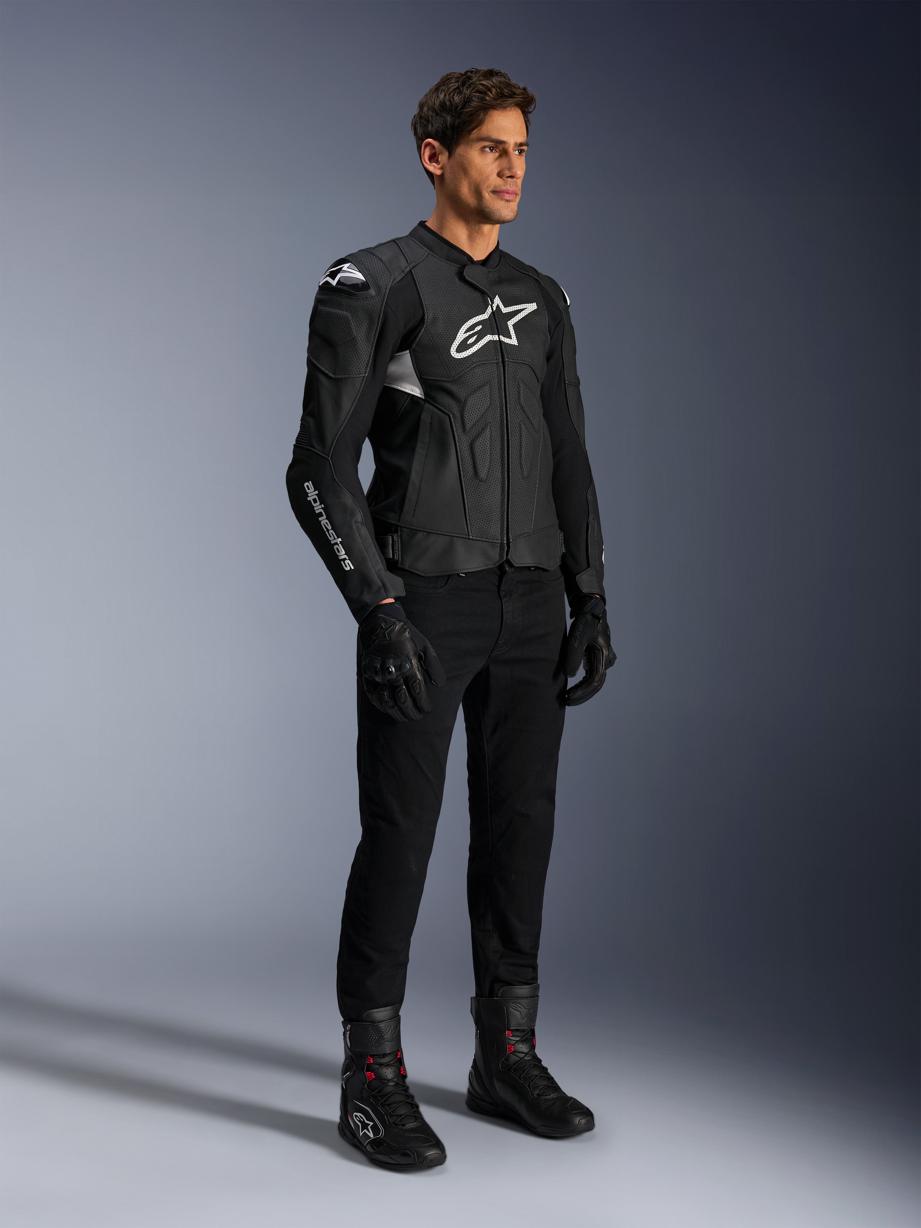 Alpinestars Dusk Airflow Leather Jacket, czarno-biała kurtka skórzana, prezentowana z czarnymi spodniami motocyklowymi, rękawicami i butami, posiada perforowane panele skórzane i białe logo, sportowy styl wyścigowy.