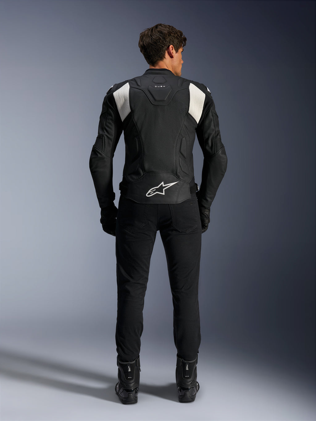Alpinestars Dusk Airflow Leather Jacket, czarno-biała, widok z tyłu motocyklisty w czarnych spodniach i butach, z perforowaną skórą dla wentylacji, białymi elastycznymi panelami akordeonowymi na ramionach i białym logo w dolnej części pleców