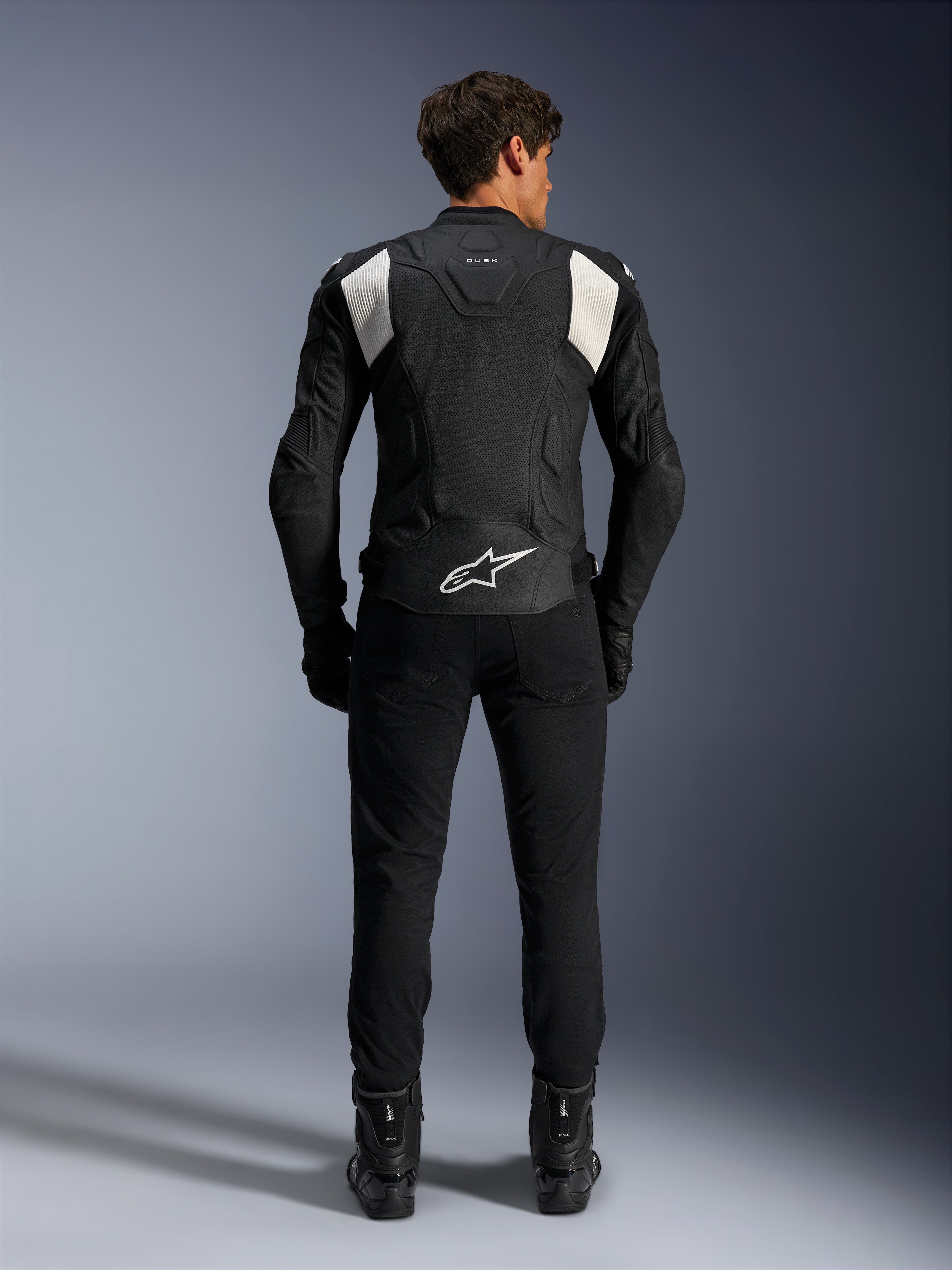 Alpinestars Dusk Airflow Leather Jacket, czarno-biała, widok z tyłu motocyklisty w czarnych spodniach i butach, z perforowaną skórą dla wentylacji, białymi elastycznymi panelami akordeonowymi na ramionach i białym logo w dolnej części pleców