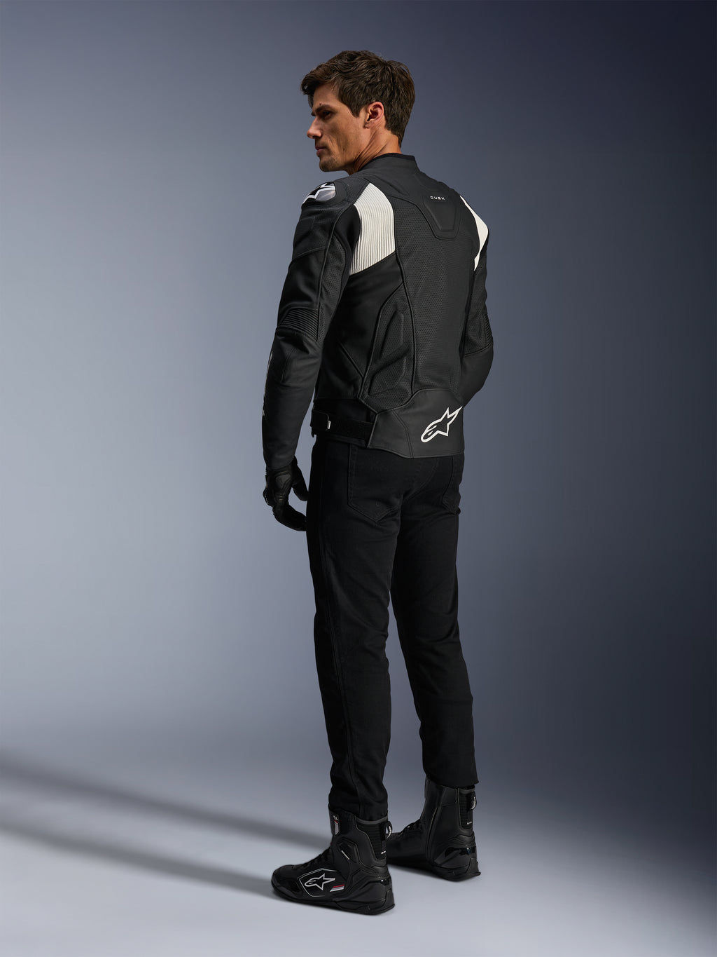 Alpinestars Dusk Airflow Leather Jacket, czarno-biała, widok z tyłu na modela ubranego w perforowaną kurtkę skórzaną z dopasowanymi czarnymi spodniami i butami, białe akordeonowe panele elastyczne i wyraźne logo w dolnej części pleców, sportowy strój motocyklowy