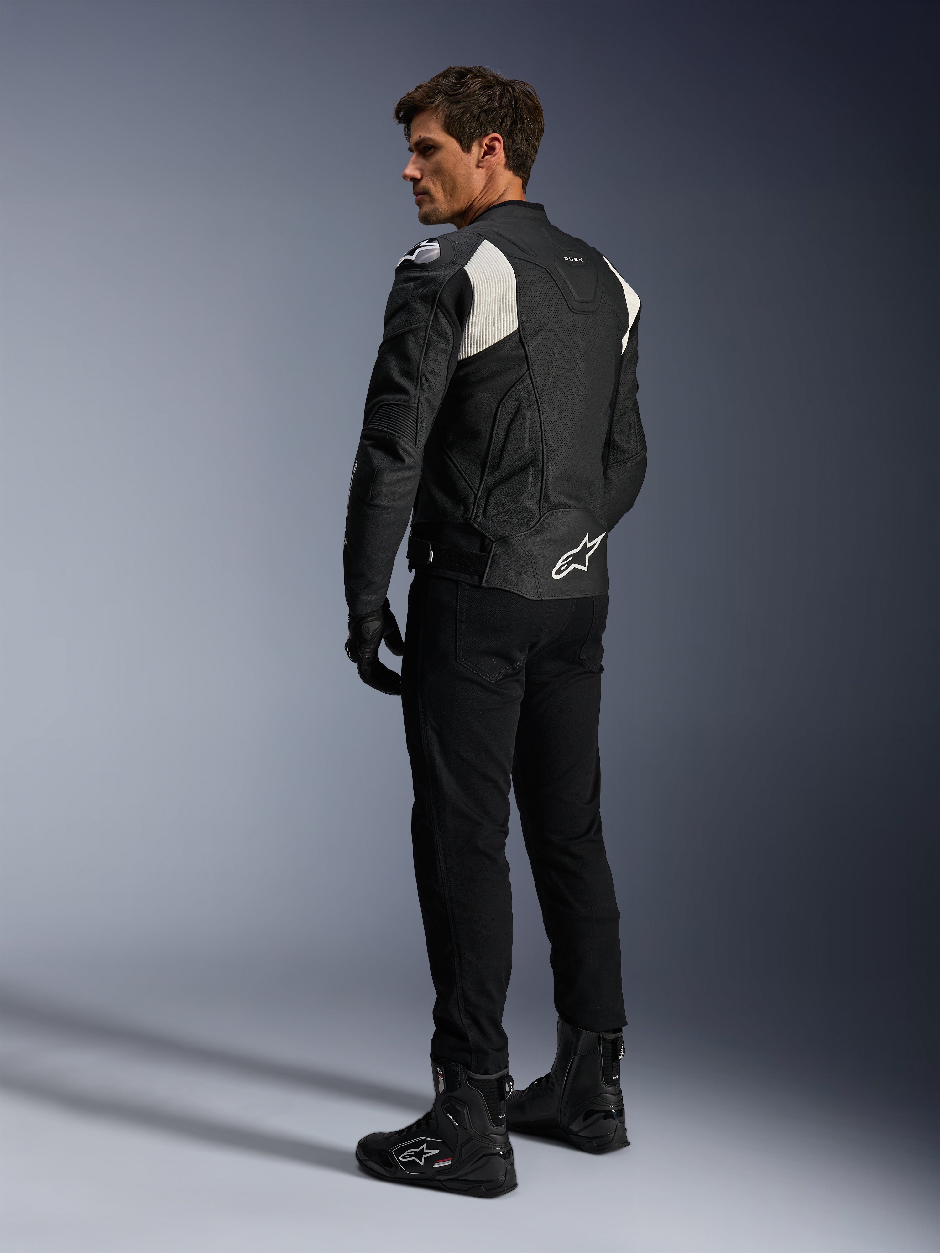 Alpinestars Dusk Airflow Leather Jacket, czarno-biała, widok z tyłu na modela ubranego w perforowaną kurtkę skórzaną z dopasowanymi czarnymi spodniami i butami, białe akordeonowe panele elastyczne i wyraźne logo w dolnej części pleców, sportowy strój motocyklowy