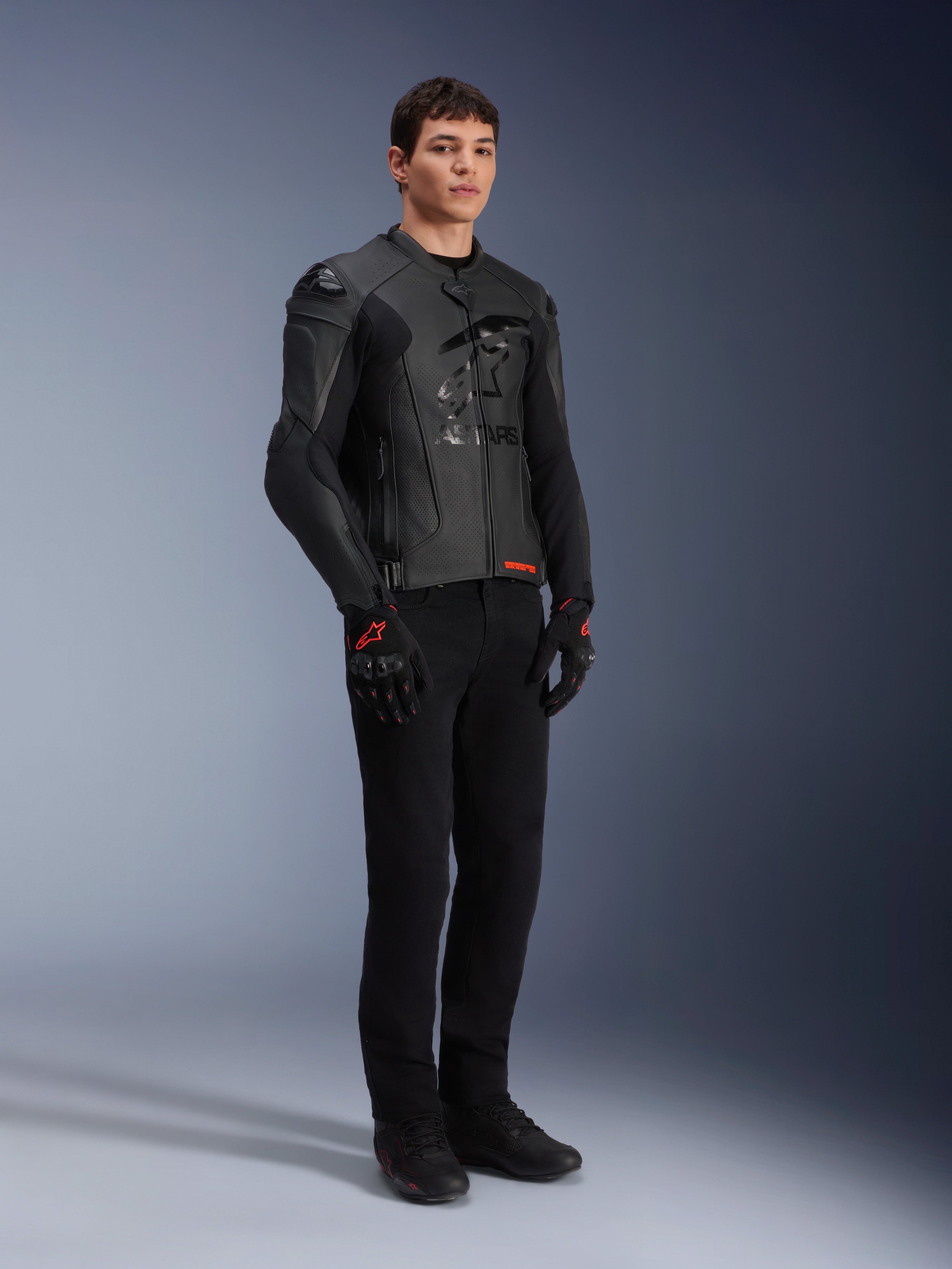 Skórzana kurtka Alpinestars GP Plus R V4 Airflow, czarna, prezentowana przez modela w czarnych spodniach motocyklowych i rękawicach z czerwonymi akcentami, perforowane panele skórzane, zintegrowane slidery ramion, ujęcie studyjne całej sylwetki.