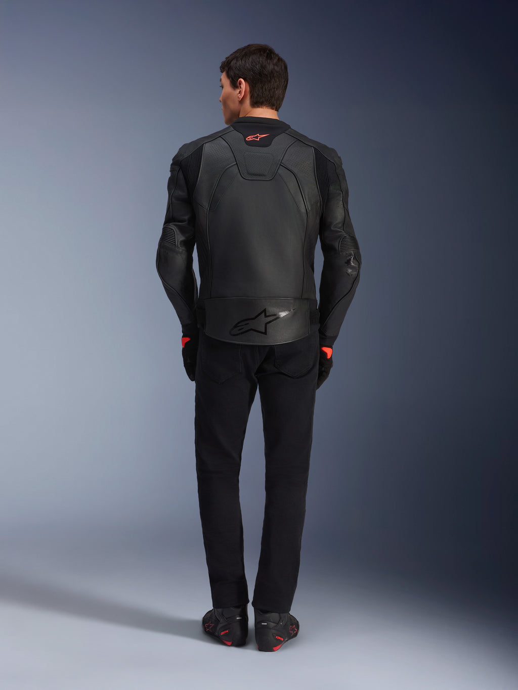 Skórzana kurtka Alpinestars GP Plus R V4 Airflow, czarna, widok od tyłu na motocyklistę w pełnym ekwipunku, posiada perforowaną skórę dla wentylacji i czerwone akcenty z logo, smukły sportowy krój, w zestawieniu z czarnymi spodniami i butami motocyklowymi