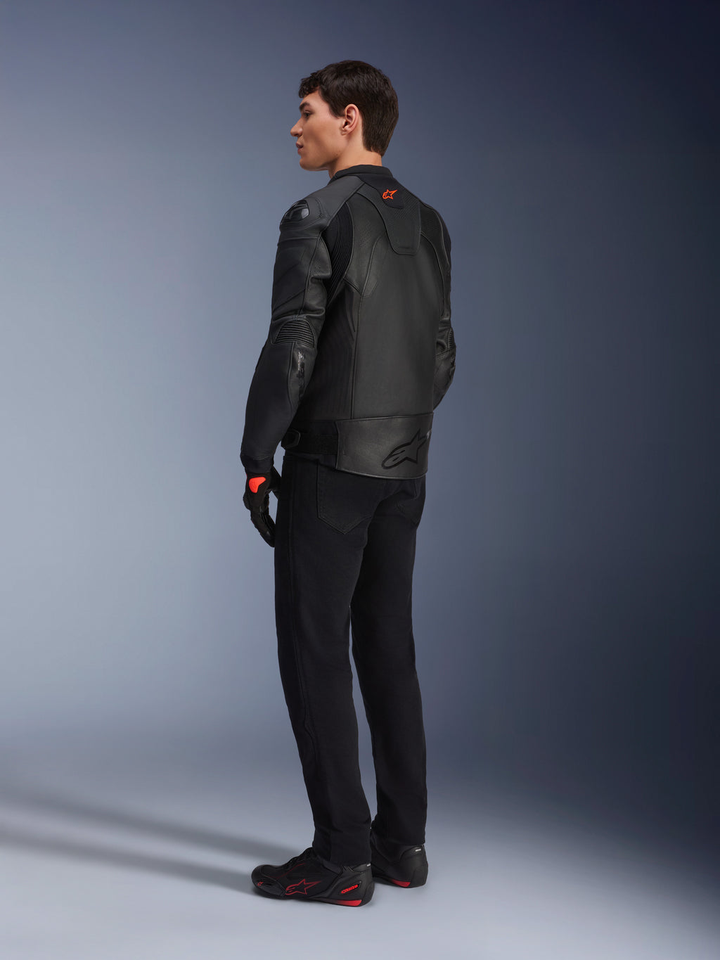 Alpinestars GP Plus R V4 Airflow Leather Jacket, męska sportowa kurtka motocyklowa, czarna, widok na modelu z tyłu pod kątem trzech czwartych, posiada perforowane panele skórzane, czerwone logo na plecach i zintegrowaną ochronę ramion, noszona z czarnymi spodniami i butami motocyklowymi