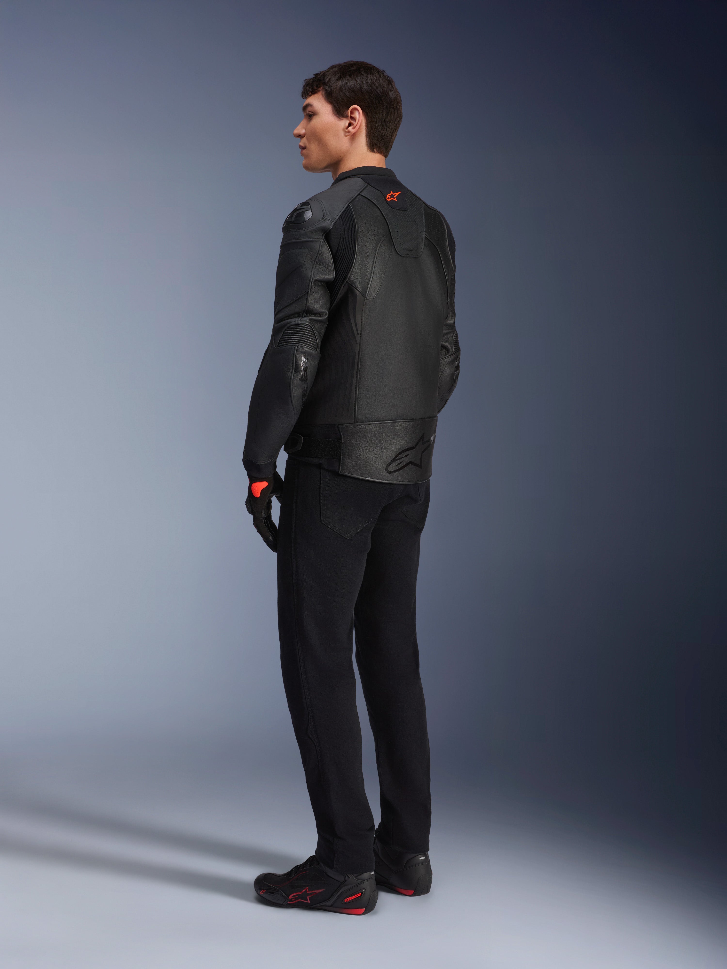 Alpinestars GP Plus R V4 Airflow Leather Jacket, męska sportowa kurtka motocyklowa, czarna, widok na modelu z tyłu pod kątem trzech czwartych, posiada perforowane panele skórzane, czerwone logo na plecach i zintegrowaną ochronę ramion, noszona z czarnymi spodniami i butami motocyklowymi