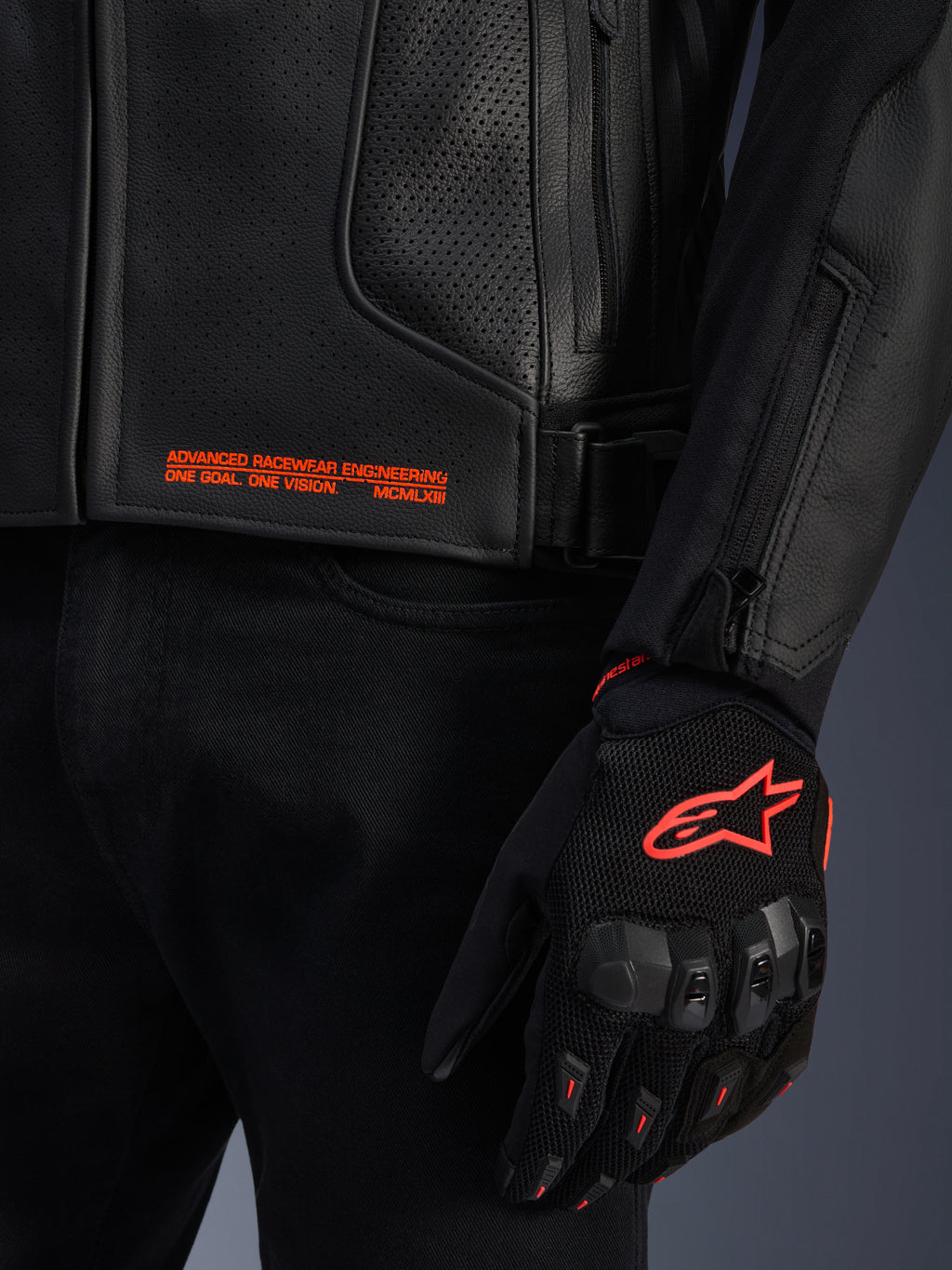 Skórzana kurtka Alpinestars GP Plus R V4 Airflow, zbliżenie na czarną perforowaną skórę z pomarańczowym napisem graficznym, zestawiona z czarno-pomarańczową rękawicą motocyklową z logo Alpinestars