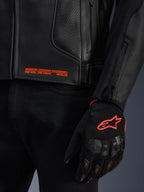 Skórzana kurtka Alpinestars GP Plus R V4 Airflow, zbliżenie na czarną perforowaną skórę z pomarańczowym napisem graficznym, zestawiona z czarno-pomarańczową rękawicą motocyklową z logo Alpinestars