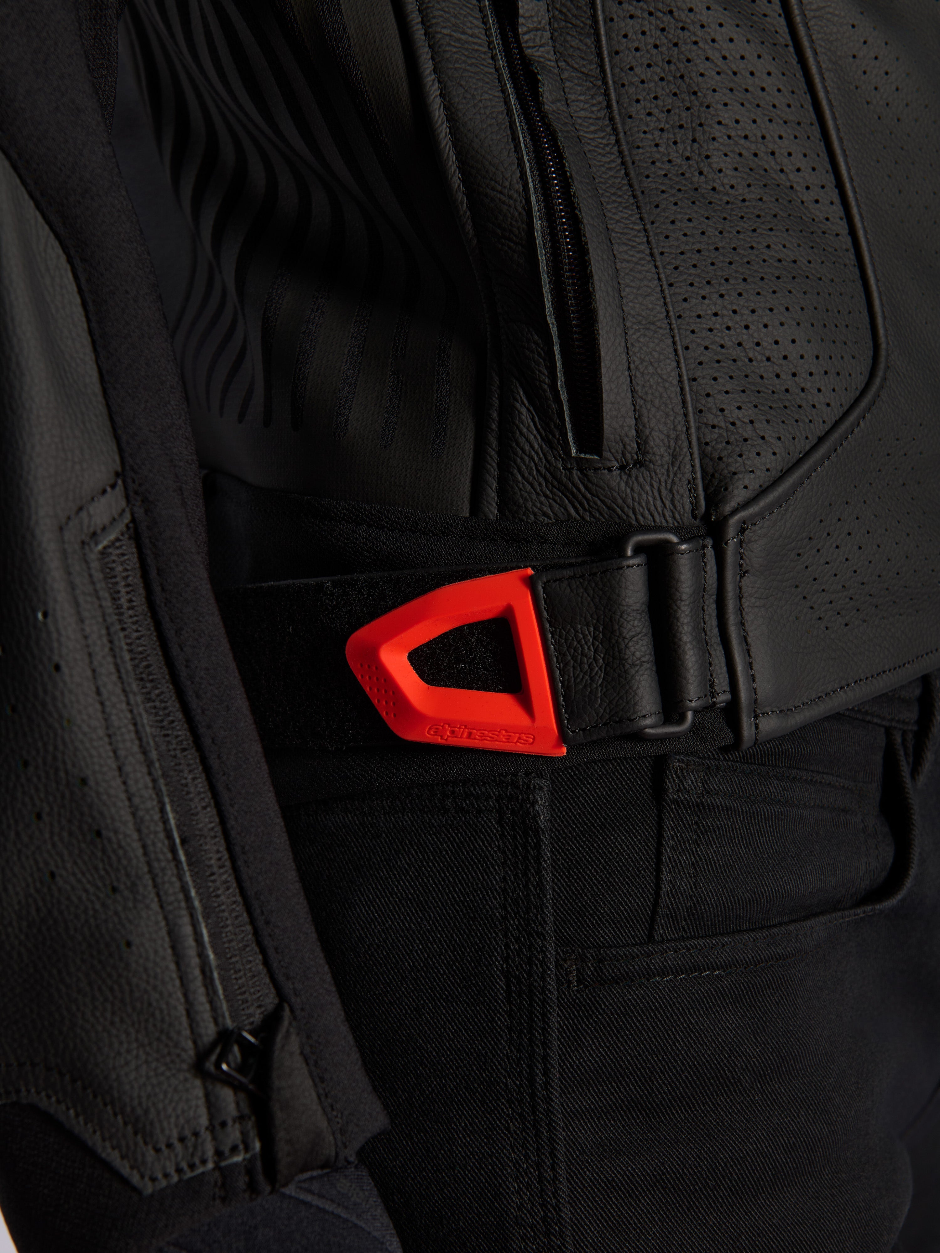 Alpinestars GP Plus R V4 Airflow Leather Jacket close-up, black perforated leather with a vibrant red fluo hook-and-loop waist adjustment strap, featuring a zippered side pocket and ergonomic textured panels.