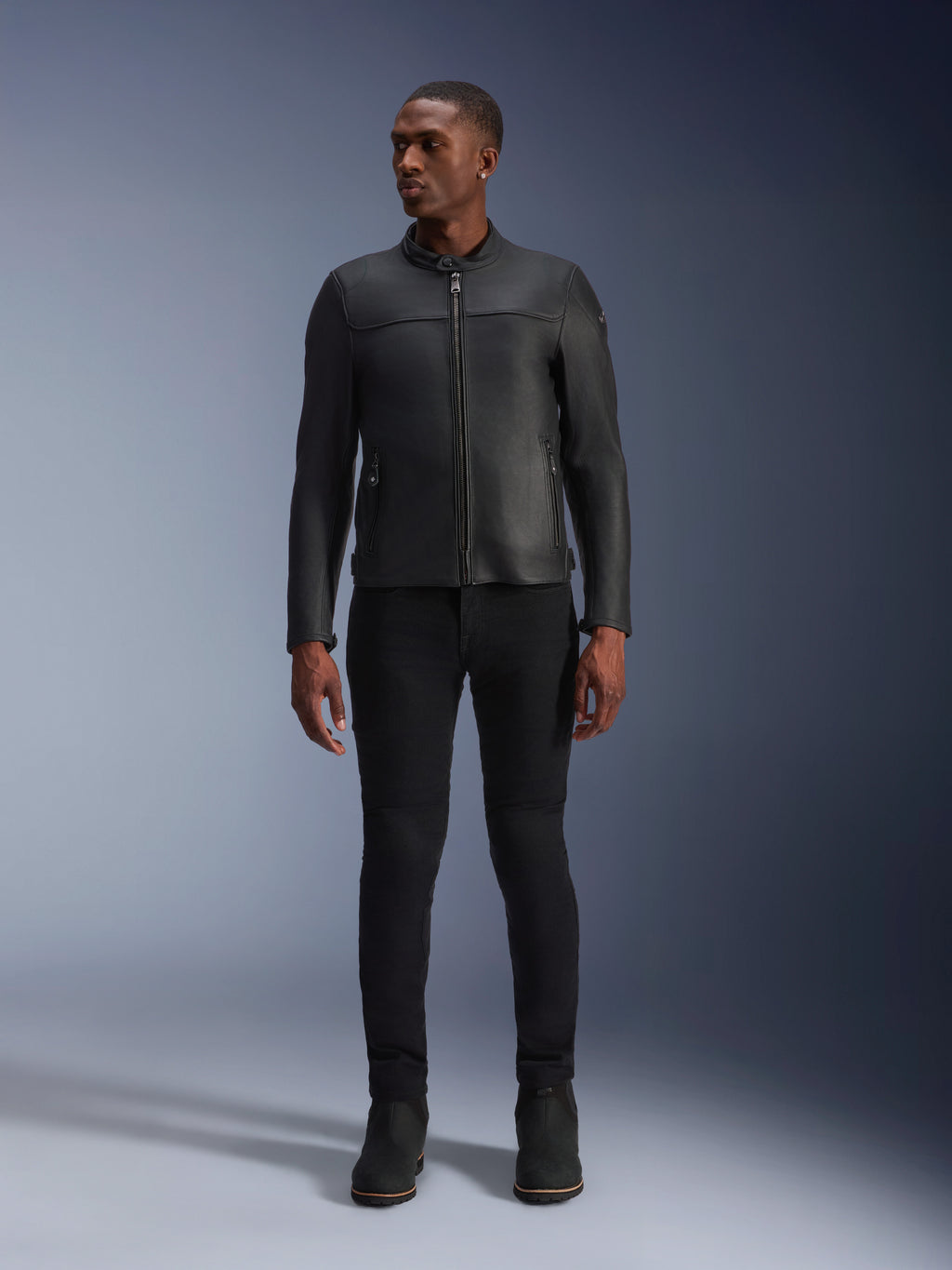 Męski model w skórzanej kurtce Alpinestars Blacktrack, czarna skóra bydlęca premium, nowoczesny minimalistyczny design motocyklowy o dopasowanym kroju, zestawiony z czarnymi spodniami motocyklowymi i butami, miejski styl współczesny