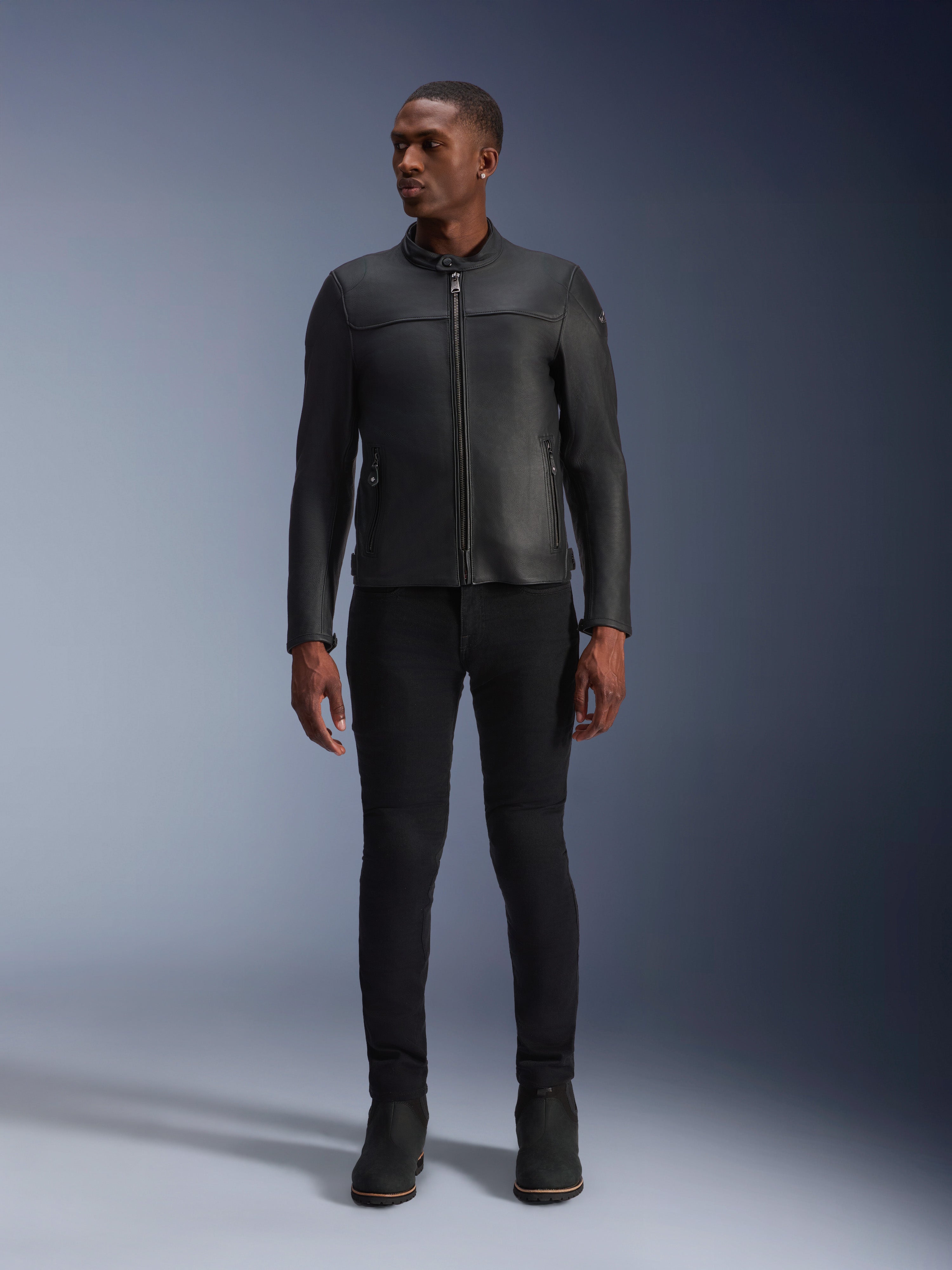 Męski model w skórzanej kurtce Alpinestars Blacktrack, czarna skóra bydlęca premium, nowoczesny minimalistyczny design motocyklowy o dopasowanym kroju, zestawiony z czarnymi spodniami motocyklowymi i butami, miejski styl współczesny