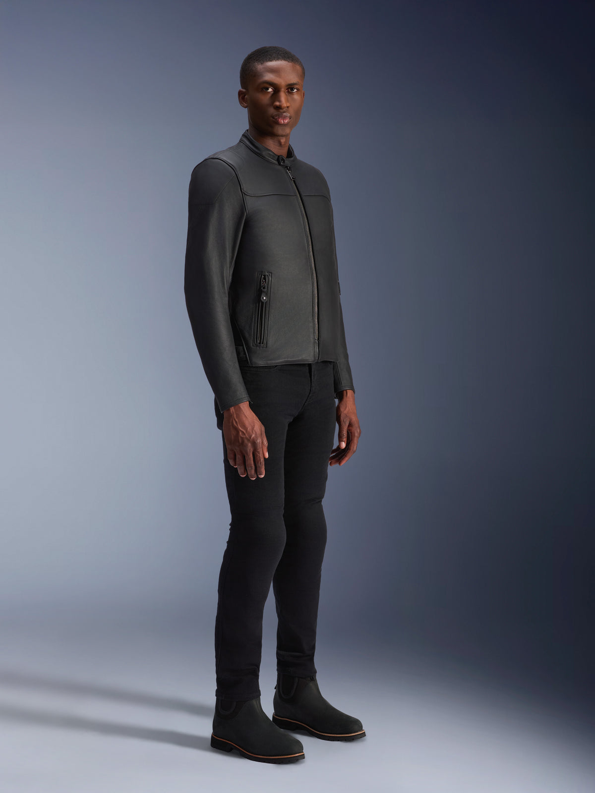Męski model w skórzanej kurtce Alpinestars Blacktrack, czarna (Black on Black), nowoczesny styl motocyklowy z wysokiej jakości skóry i metalowymi detalami zamków, zestawiona z czarnymi spodniami i butami, nowoczesny miejski wygląd