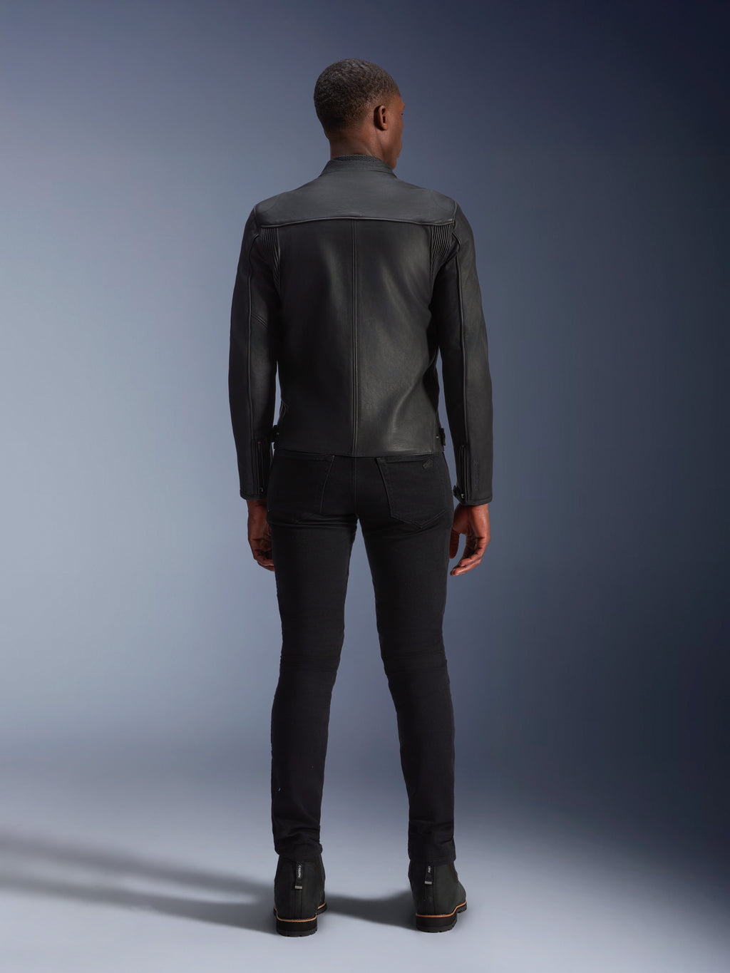 Skórzana kurtka Alpinestars Blacktrack, widok z tyłu, czarna skóra bydlęca premium, panele akordeonowe na łopatkach zapewniające swobodę ruchów, czarne spodnie o kroju slim-fit i czarne buty, miejska współczesna odzież motocyklowa