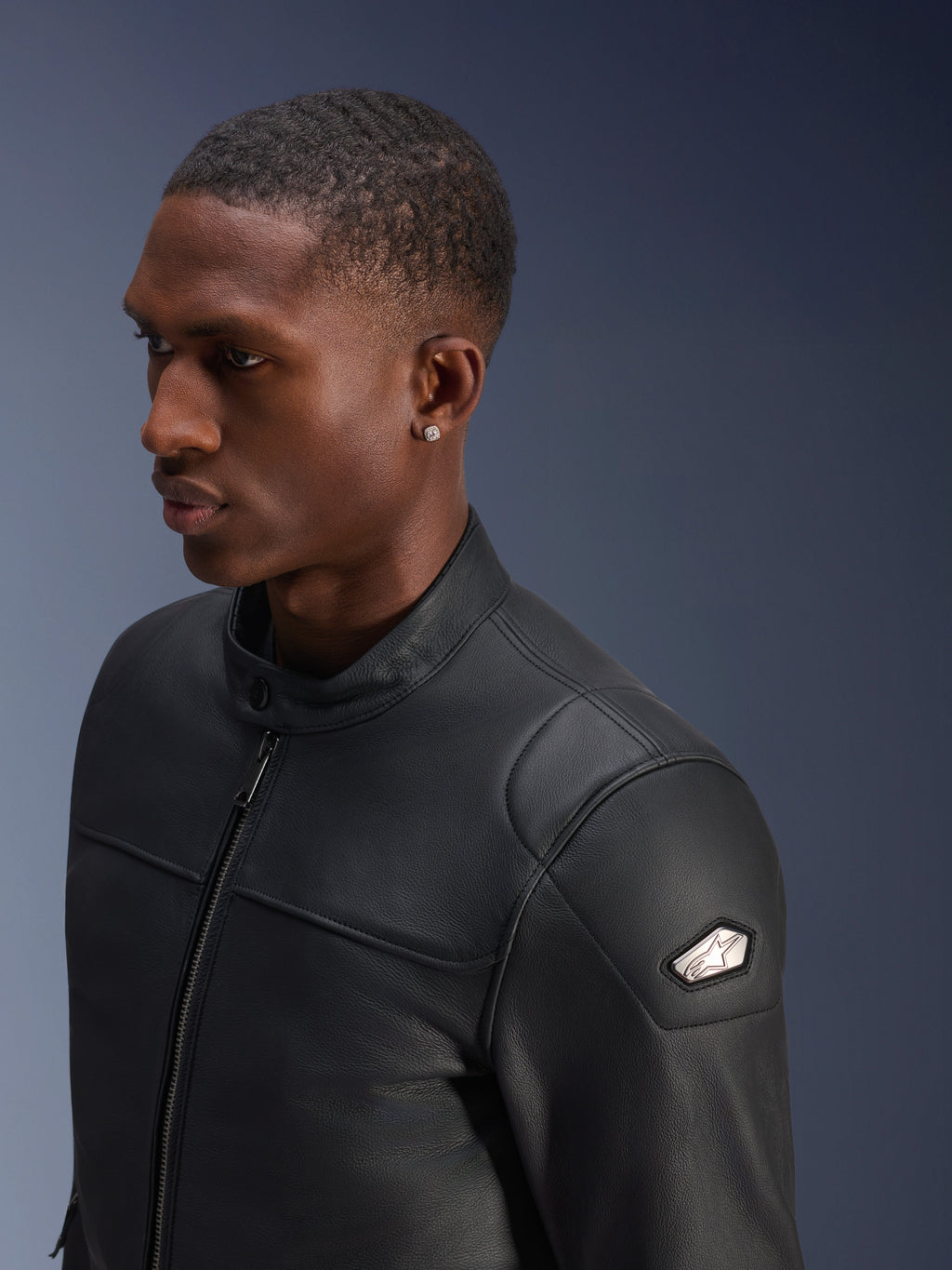 Alpinestars Blacktrack Leather Jacket, wysokiej jakości czarna skóra bydlęca, zbliżenie z profilu na mężczyznę w kurtce, metalowy zamek z przodu w kolorze gunmetal, wysoki kołnierz zapinany na guziki, metalowa naszywka z logo na ramieniu, elegancka dopasowana kurtka motocyklowa