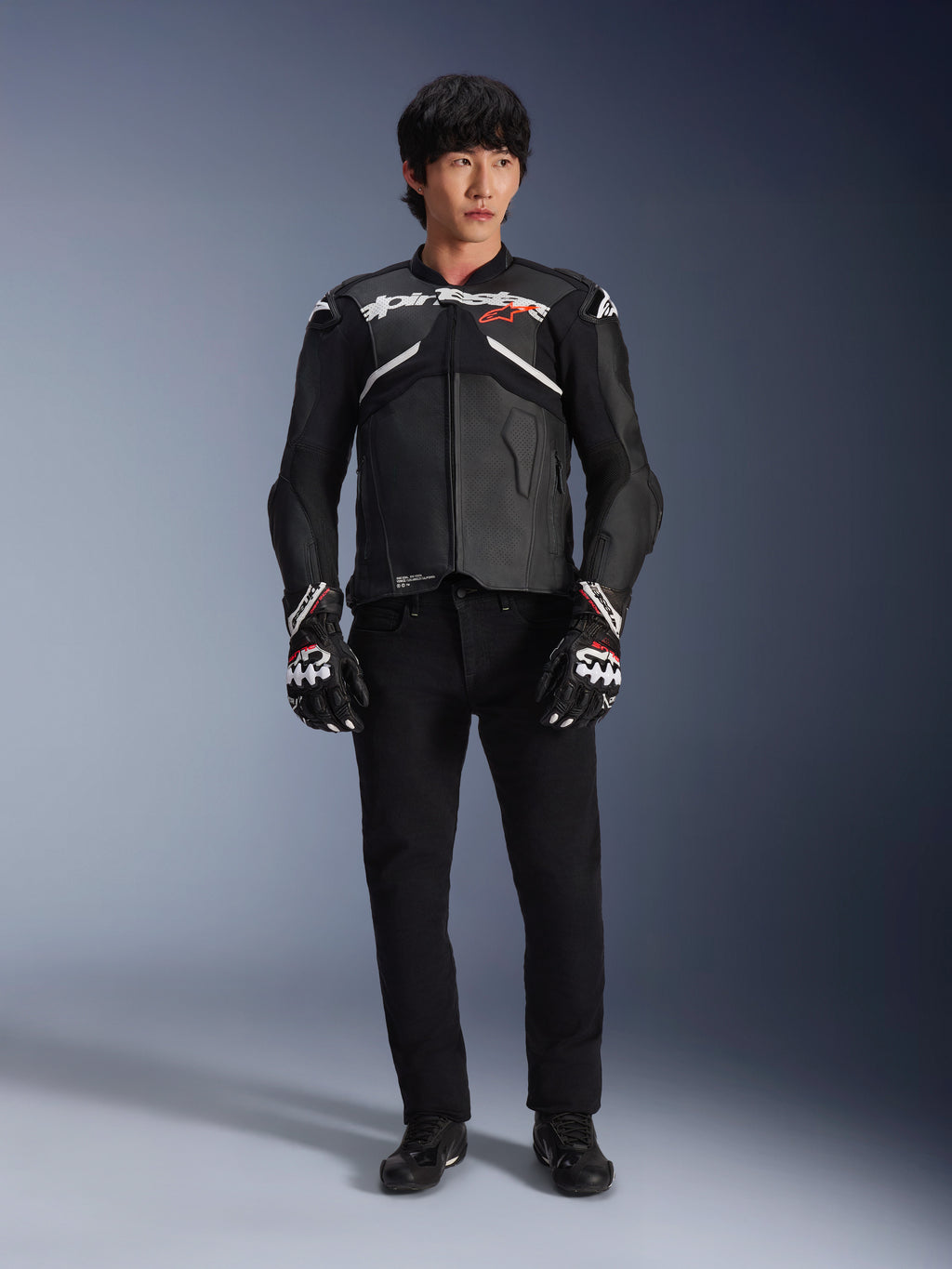 Model w skórzanej kurtce Alpinestars Atem V5 w kolorze czarno-białym, z białym brandingiem Alpinestars i czerwonym logo gwiazdy na piersi, w zestawieniu z czarnymi spodniami i czarno-białymi rękawicami wyścigowymi, profesjonalna odzież motocyklowa