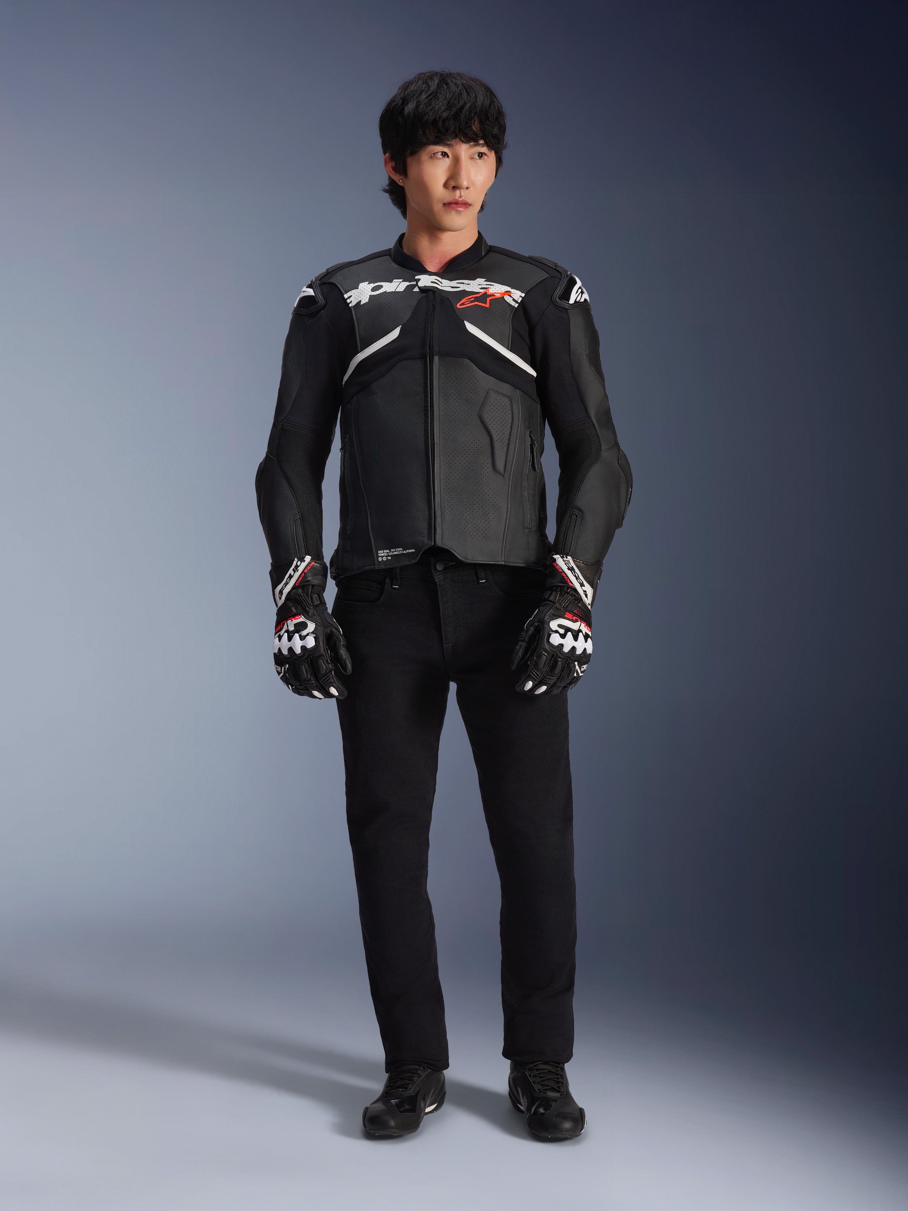 Model w skórzanej kurtce Alpinestars Atem V5 w kolorze czarno-białym, z białym brandingiem Alpinestars i czerwonym logo gwiazdy na piersi, w zestawieniu z czarnymi spodniami i czarno-białymi rękawicami wyścigowymi, profesjonalna odzież motocyklowa