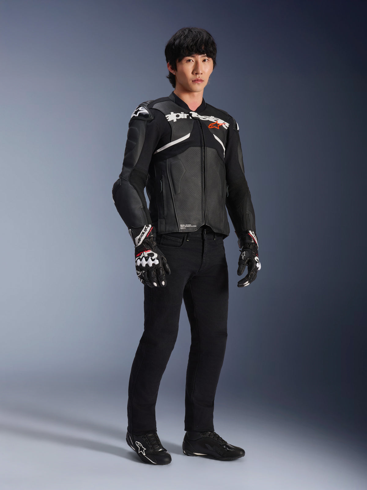 Skórzana kurtka Alpinestars Atem V5, motocyklowa kurtka wyścigowa, czarno-biała, noszona z czarnymi spodniami i dopasowanymi rękawicami, z białym logo Alpinestars na klatce piersiowej i ochronnymi protektorami ramion
