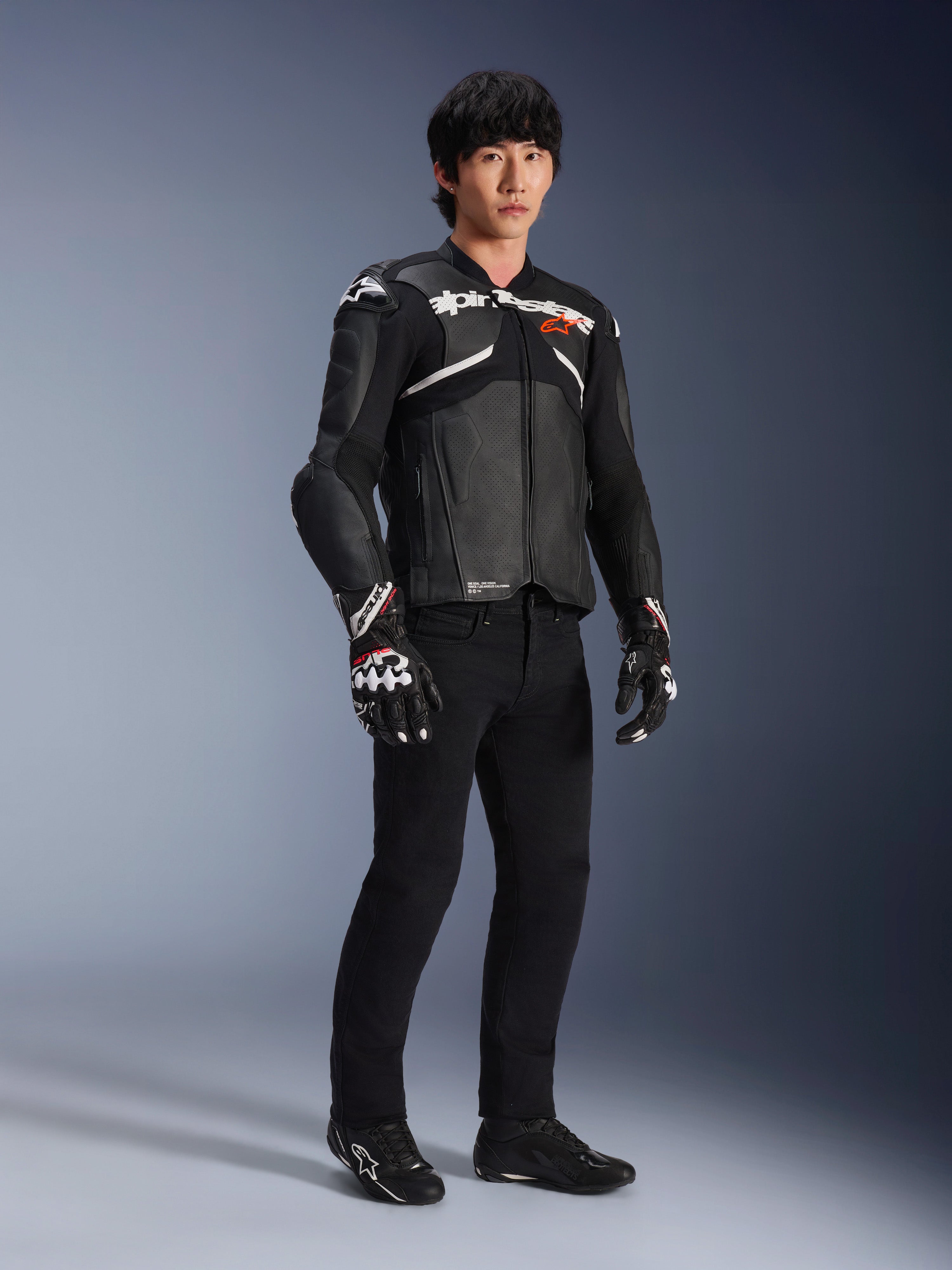 Skórzana kurtka Alpinestars Atem V5, motocyklowa kurtka wyścigowa, czarno-biała, noszona z czarnymi spodniami i dopasowanymi rękawicami, z białym logo Alpinestars na klatce piersiowej i ochronnymi protektorami ramion