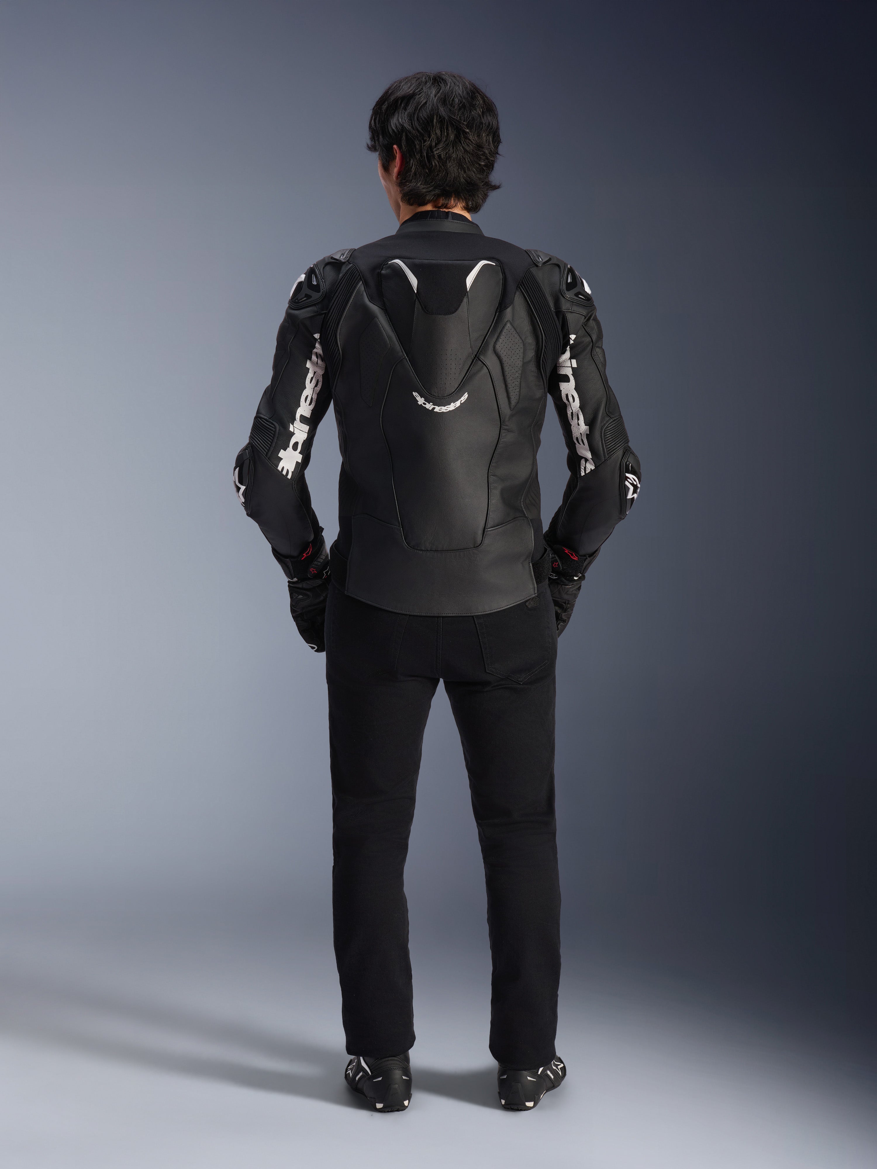 Alpinestars Atem V5 Leather Jacket, motocyklowa kurtka wyścigowa, czarno-biała, widok od tyłu na motocykliście, wyposażona w aerodynamiczny garb i białe logo Alpinestars na rękawach i plecach, techniczna skórzana konstrukcja, noszona z czarnymi spodniami i butami motocyklowymi