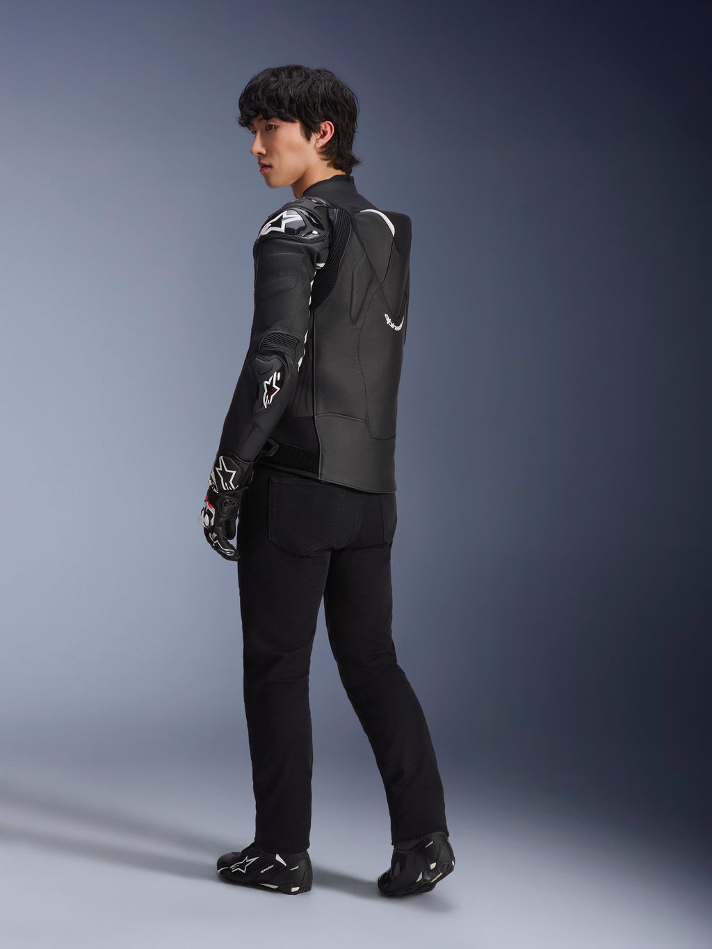 Męski model w skórzanej kurtce Alpinestars Atem V5 w kolorze czarno-białym, widok z tyłu z profilu pokazujący aerodynamiczny garb i ochraniacze DFS RR, stylizacja z czarnymi spodniami, rękawicami wyścigowymi i butami, profesjonalna wyścigowa odzież motocyklowa