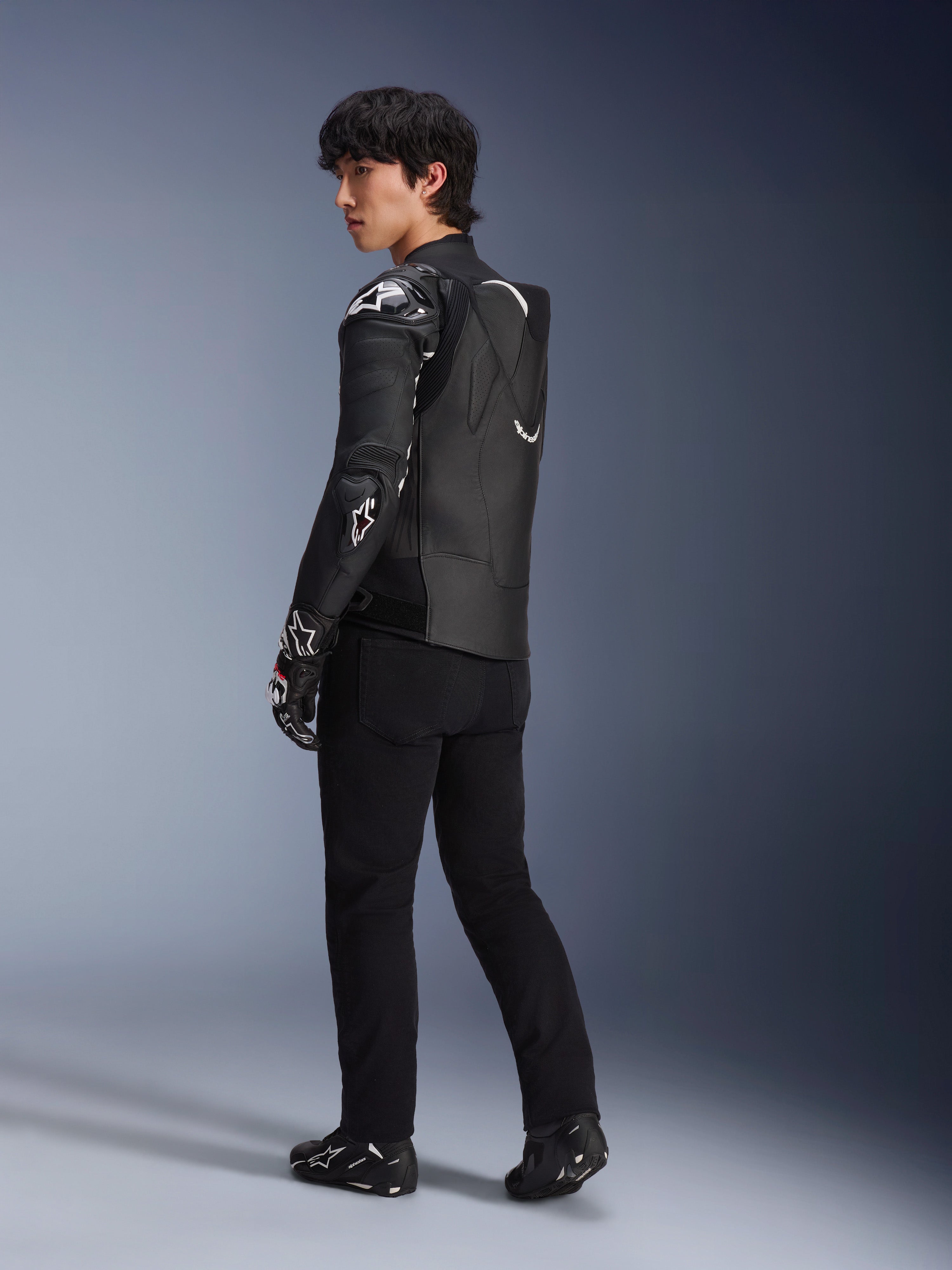 Męski model w skórzanej kurtce Alpinestars Atem V5 w kolorze czarno-białym, widok z tyłu z profilu pokazujący aerodynamiczny garb i ochraniacze DFS RR, stylizacja z czarnymi spodniami, rękawicami wyścigowymi i butami, profesjonalna wyścigowa odzież motocyklowa