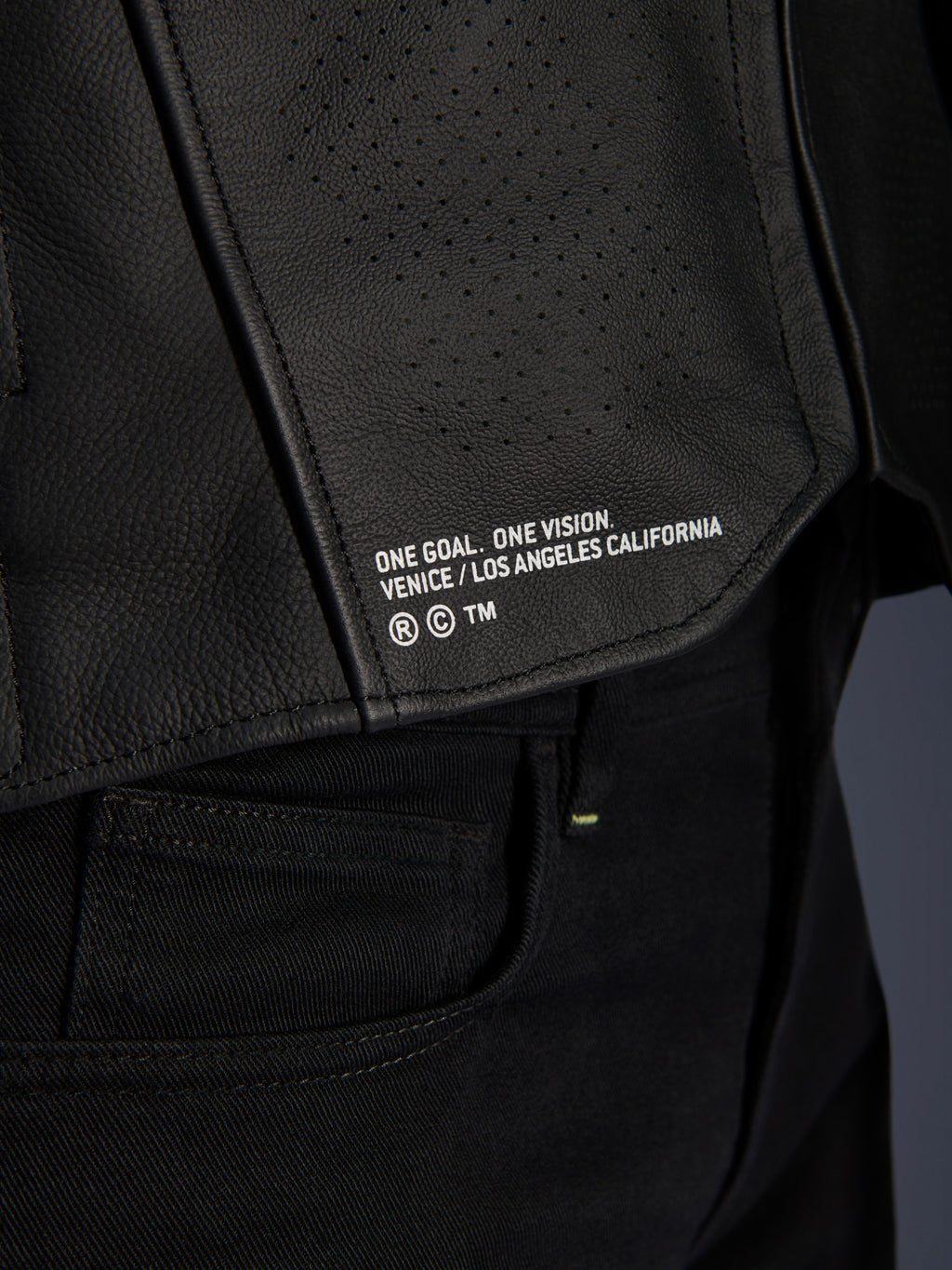 Skórzana kurtka Alpinestars Atem V5, czarna, zbliżenie na dolny tylny panel z napisem „ONE GOAL. ONE VISION. VENICE / LOS ANGELES CALIFORNIA”, widoczna tekstura perforowanej skóry