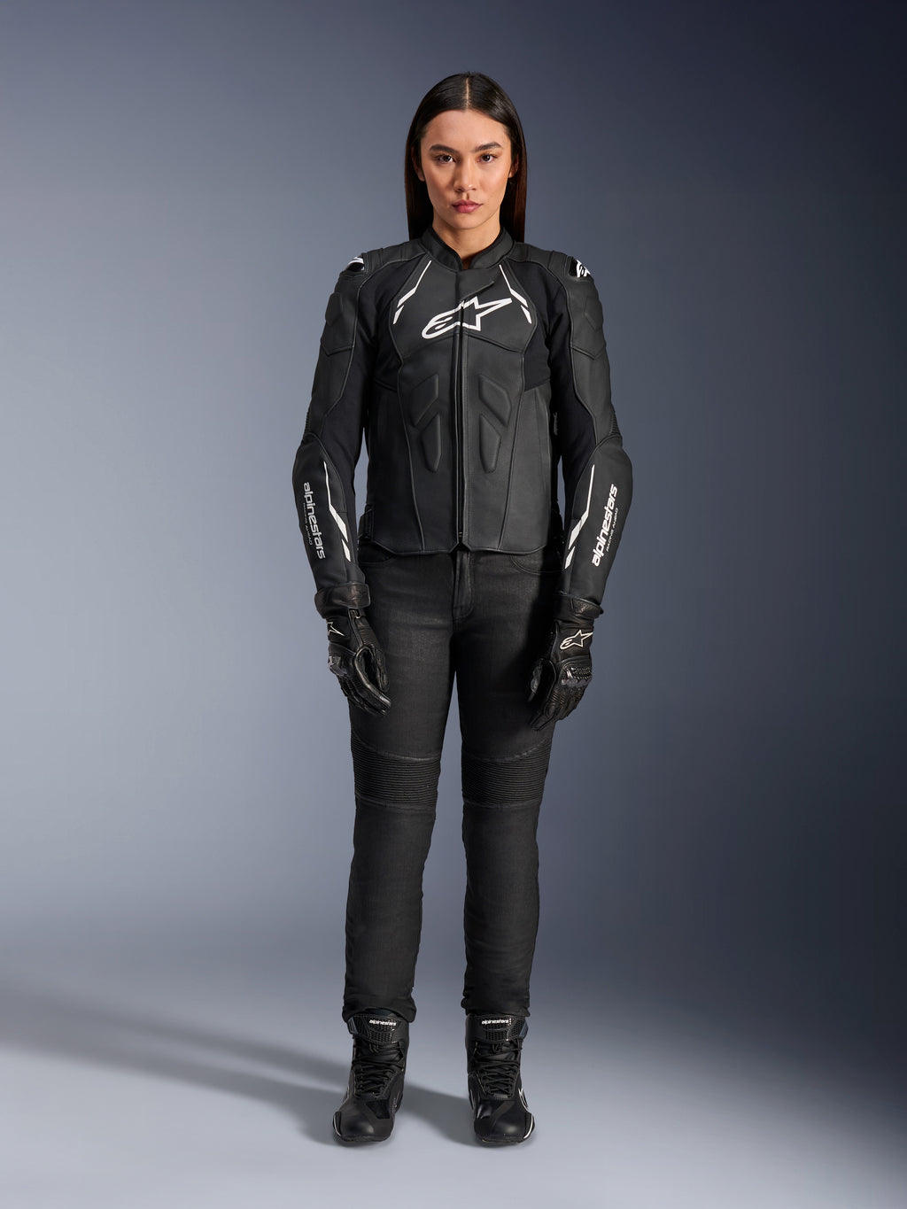 Damska skórzana kurtka Alpinestars Stella Dusk, czarno-biała, prezentowana przez modelkę z dopasowanymi czarnymi spodniami i butami, widok z przodu, z białym logo Alpinestars na klatce piersiowej i rękawach, najwyższej jakości odzież ochronna z bydlęcej skóry dla kobiet