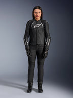 Damska skórzana kurtka Alpinestars Stella Dusk, czarno-biała, prezentowana przez modelkę z dopasowanymi czarnymi spodniami i butami, widok z przodu, z białym logo Alpinestars na klatce piersiowej i rękawach, najwyższej jakości odzież ochronna z bydlęcej skóry dla kobiet