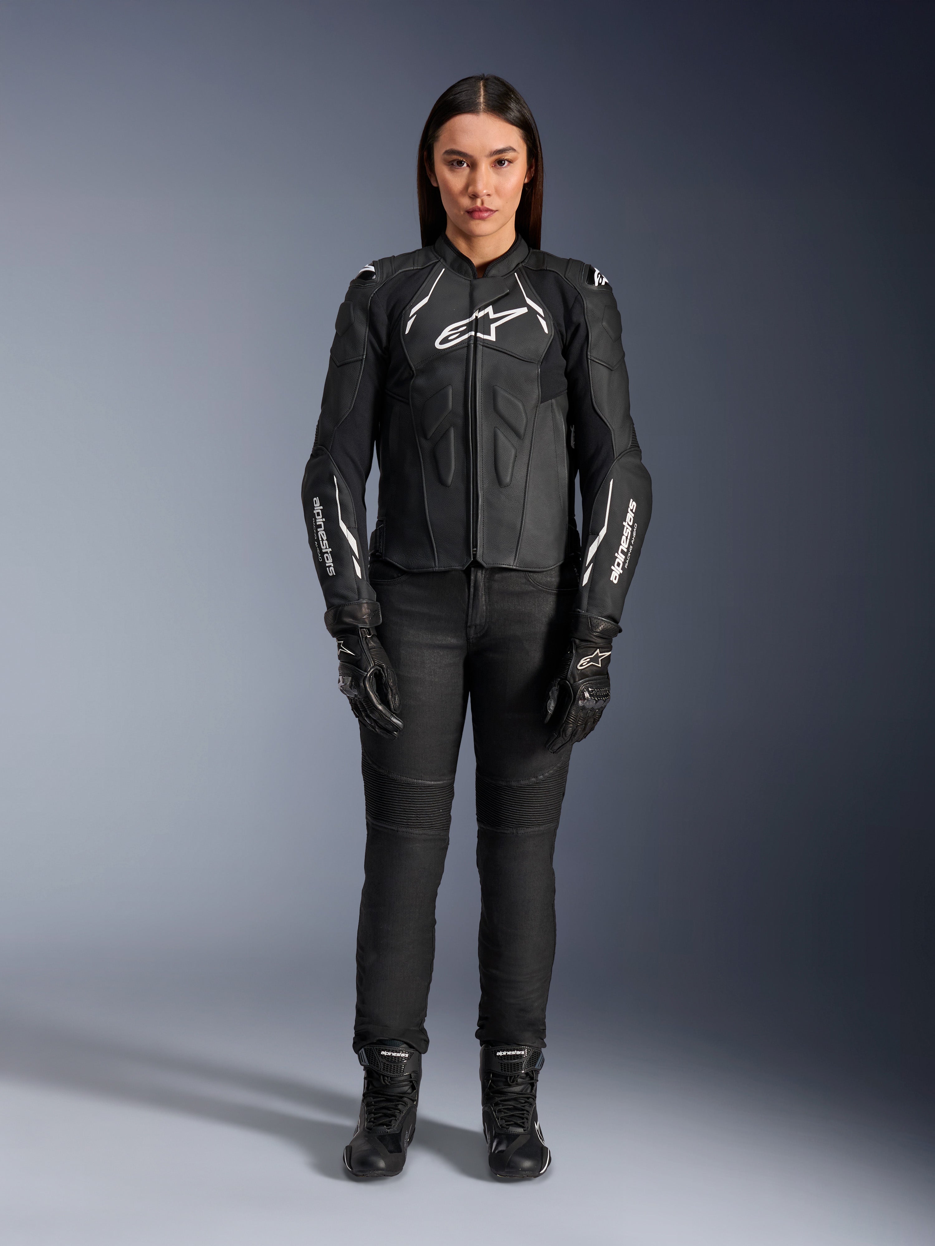 Damska skórzana kurtka Alpinestars Stella Dusk, czarno-biała, prezentowana przez modelkę z dopasowanymi czarnymi spodniami i butami, widok z przodu, z białym logo Alpinestars na klatce piersiowej i rękawach, najwyższej jakości odzież ochronna z bydlęcej skóry dla kobiet