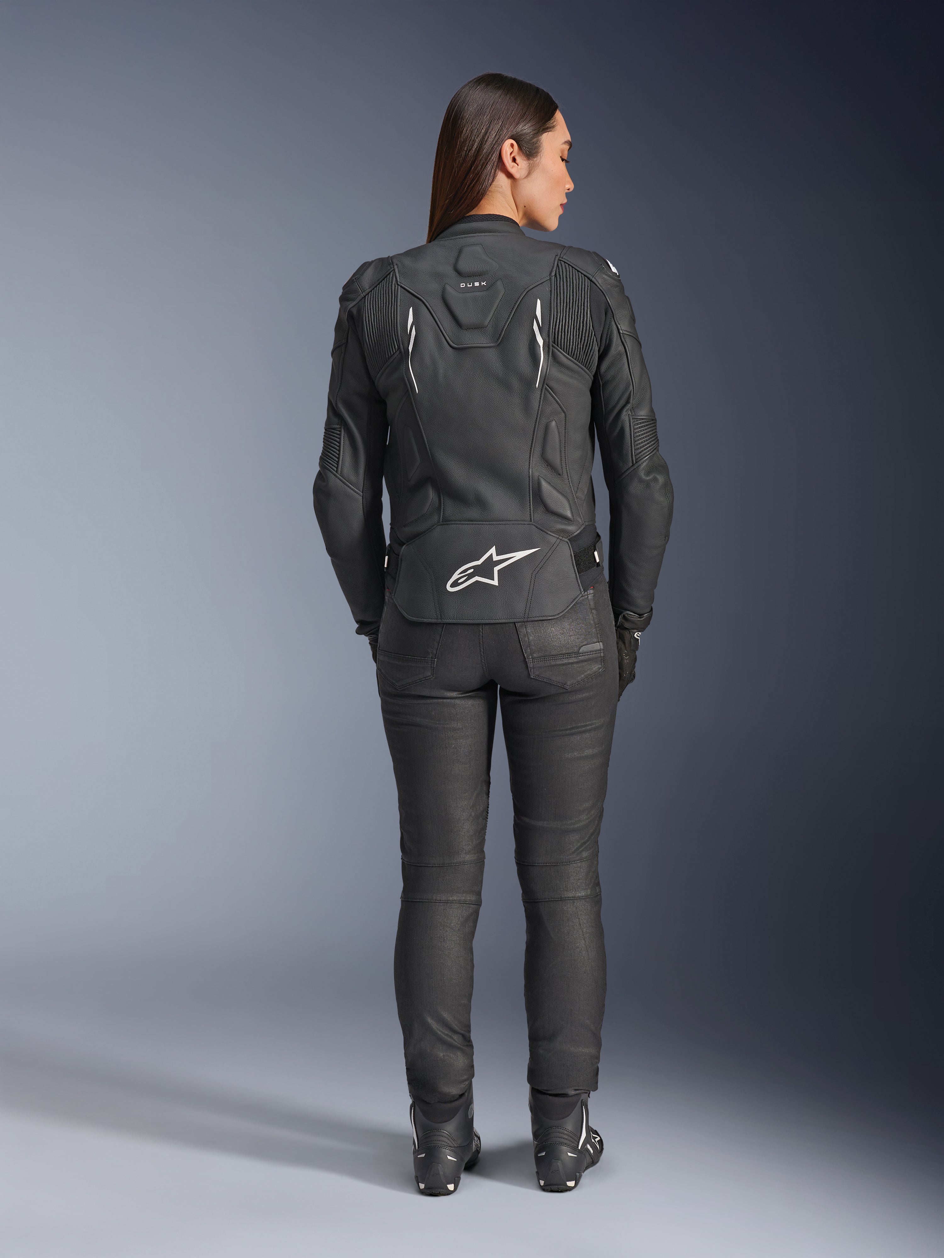Damska skórzana kurtka Alpinestars Stella Dusk, czarna, widok z tyłu z białym logo Alpinestars na dole pleców i brandingiem „DUSK” w górnej części pleców, noszona z czarnymi spodniami motocyklowymi i butami, prezentująca dopasowany damski krój oraz ochronną skórzaną konstrukcję.