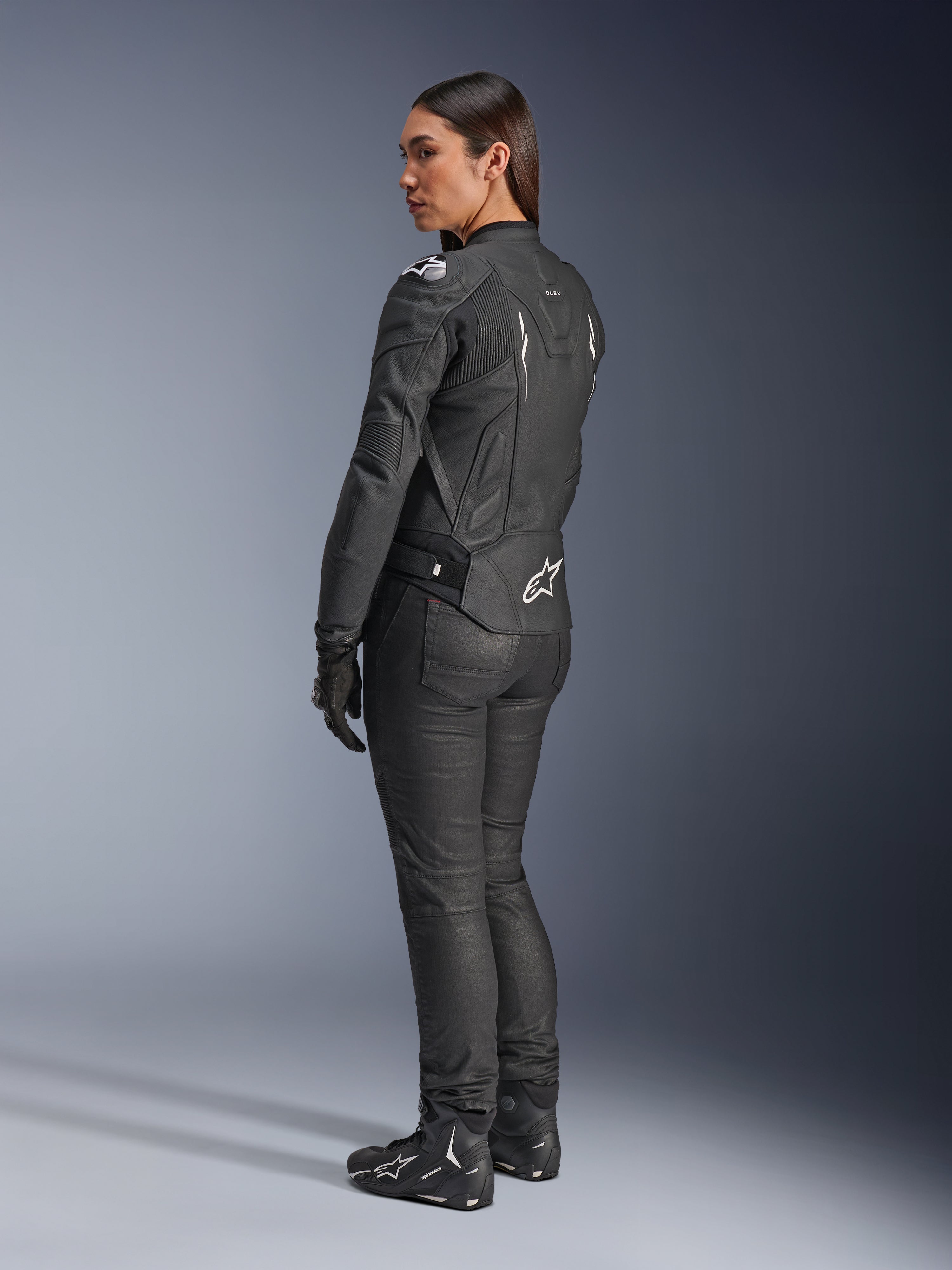 Alpinestars Stella Dusk Leather Jacket, damska kurtka motocyklowa, czarno-biała, widok z tyłu z białym logo Alpinestars na dolnej części pleców i ramionach, noszona z czarnymi spodniami i butami motocyklowymi, sportowy ergonomiczny krój do jazdy szosowej