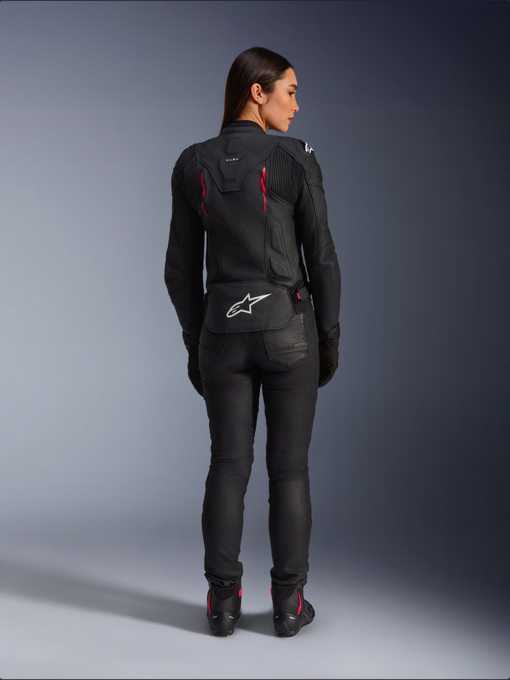 Skórzana kurtka Alpinestars Stella Dusk, widok z tyłu, czarna skóra z akcentami Diva Pink i białymi, białe logo Alpinestars w dolnej części pleców, różowe detale wentylacyjne, noszona z czarnymi spodniami i butami motocyklowymi, sportowa odzież motocyklowa premium dla kobiet