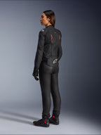 Damska skórzana kurtka Alpinestars Stella Dusk, Black Diva Pink White, widok z tyłu pod kątem 3/4 na motocyklistce, ubrana w czarne spodnie i buty motocyklowe, smukła skórzana konstrukcja z różowymi akcentami i białym logo Alpinestars na dole pleców.