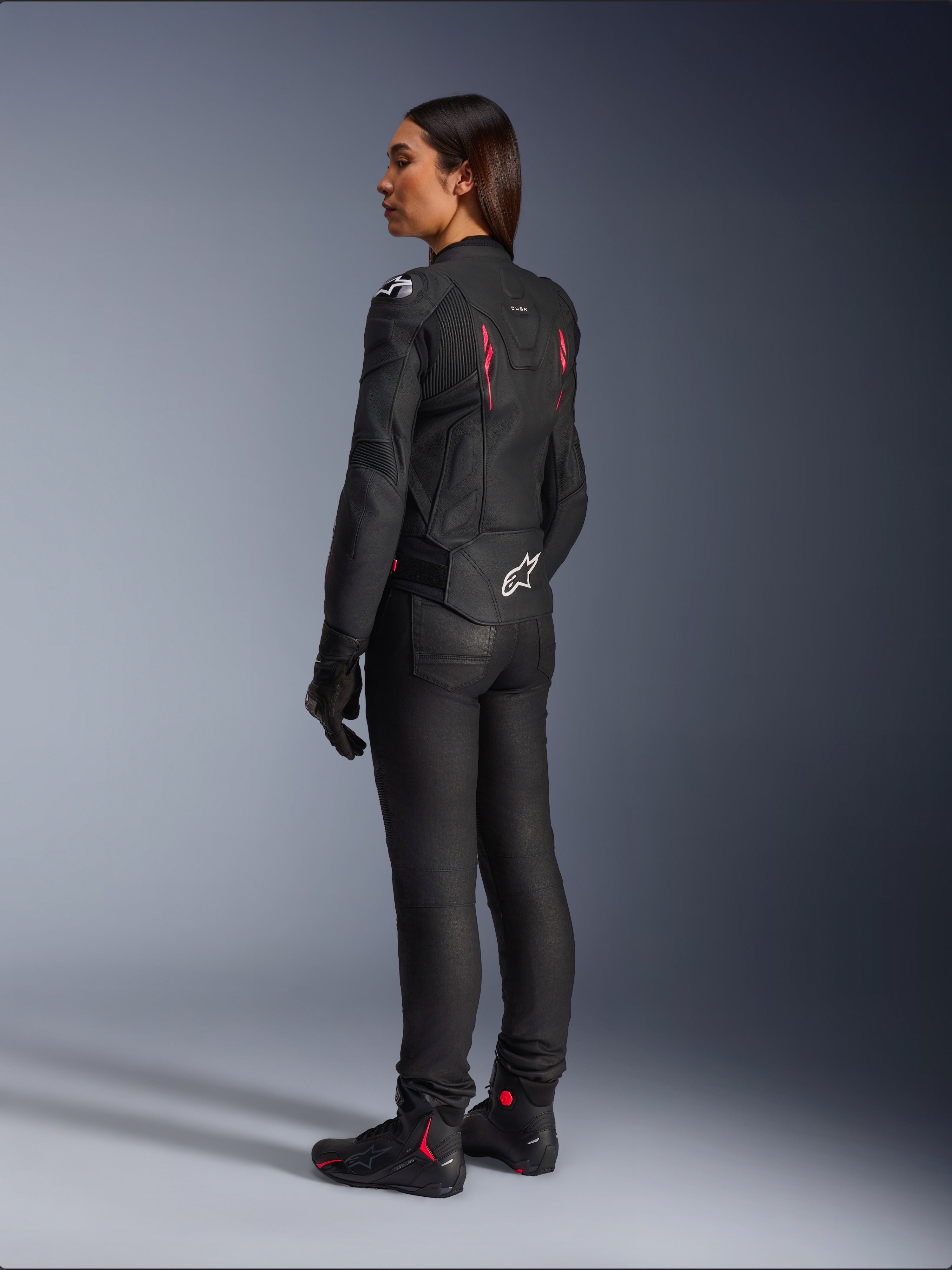 Damska skórzana kurtka Alpinestars Stella Dusk, Black Diva Pink White, widok z tyłu pod kątem 3/4 na motocyklistce, ubrana w czarne spodnie i buty motocyklowe, smukła skórzana konstrukcja z różowymi akcentami i białym logo Alpinestars na dole pleców.
