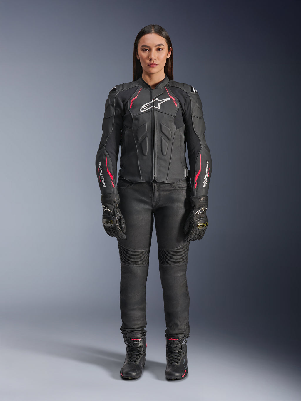 Kobieta ubrana w skórzaną kurtkę Alpinestars Stella Dusk w kolorze Black Diva Pink White, z białym logo na klatce piersiowej i różowymi akcentami na ramionach i rękawach, w zestawieniu z czarnymi spodniami motocyklowymi, rękawicami i butami.
