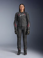 Kobieta ubrana w skórzaną kurtkę Alpinestars Stella Dusk w kolorze Black Diva Pink White, z białym logo na klatce piersiowej i różowymi akcentami na ramionach i rękawach, w zestawieniu z czarnymi spodniami motocyklowymi, rękawicami i butami.