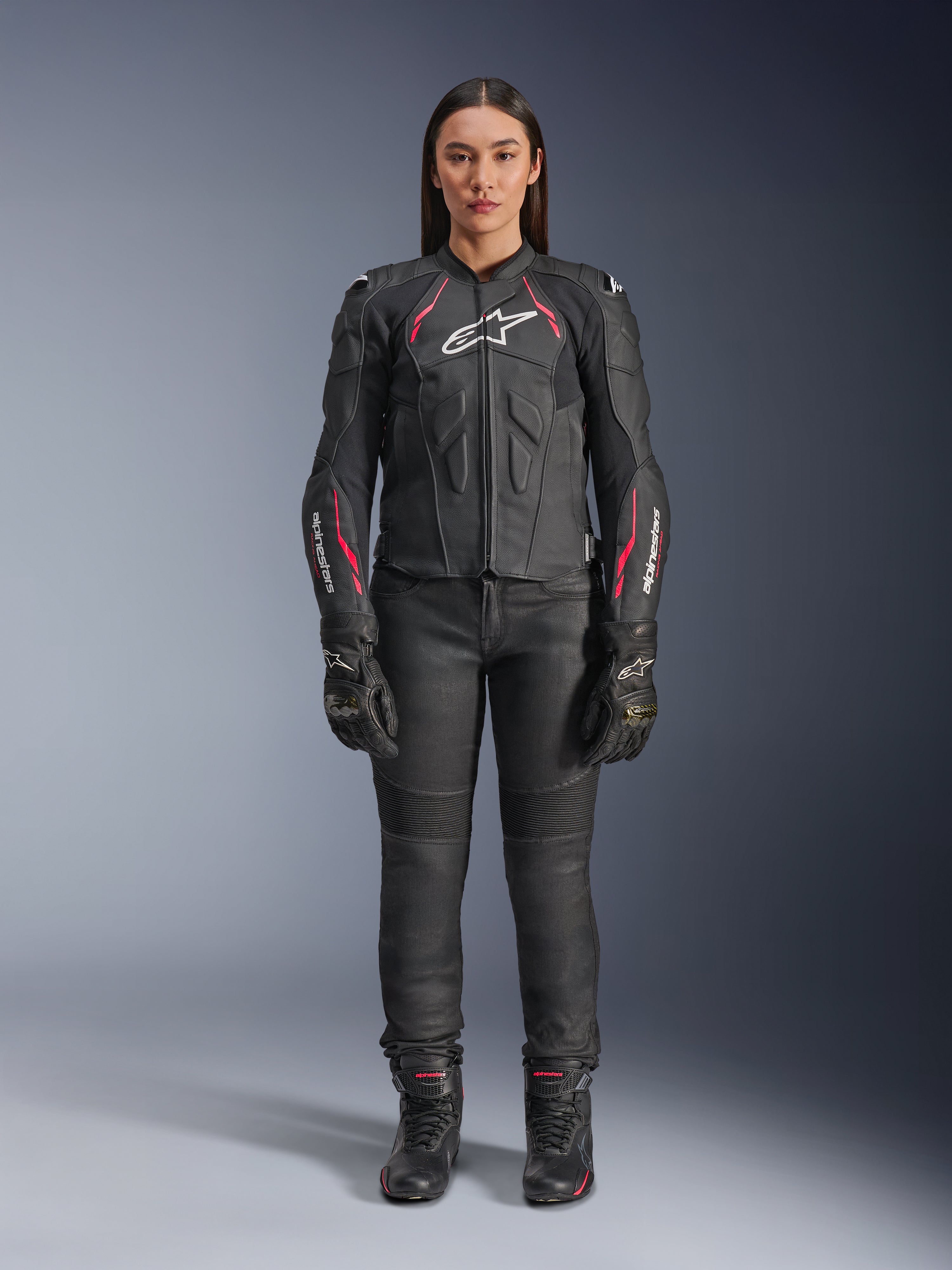 Kobieta ubrana w skórzaną kurtkę Alpinestars Stella Dusk w kolorze Black Diva Pink White, z białym logo na klatce piersiowej i różowymi akcentami na ramionach i rękawach, w zestawieniu z czarnymi spodniami motocyklowymi, rękawicami i butami.