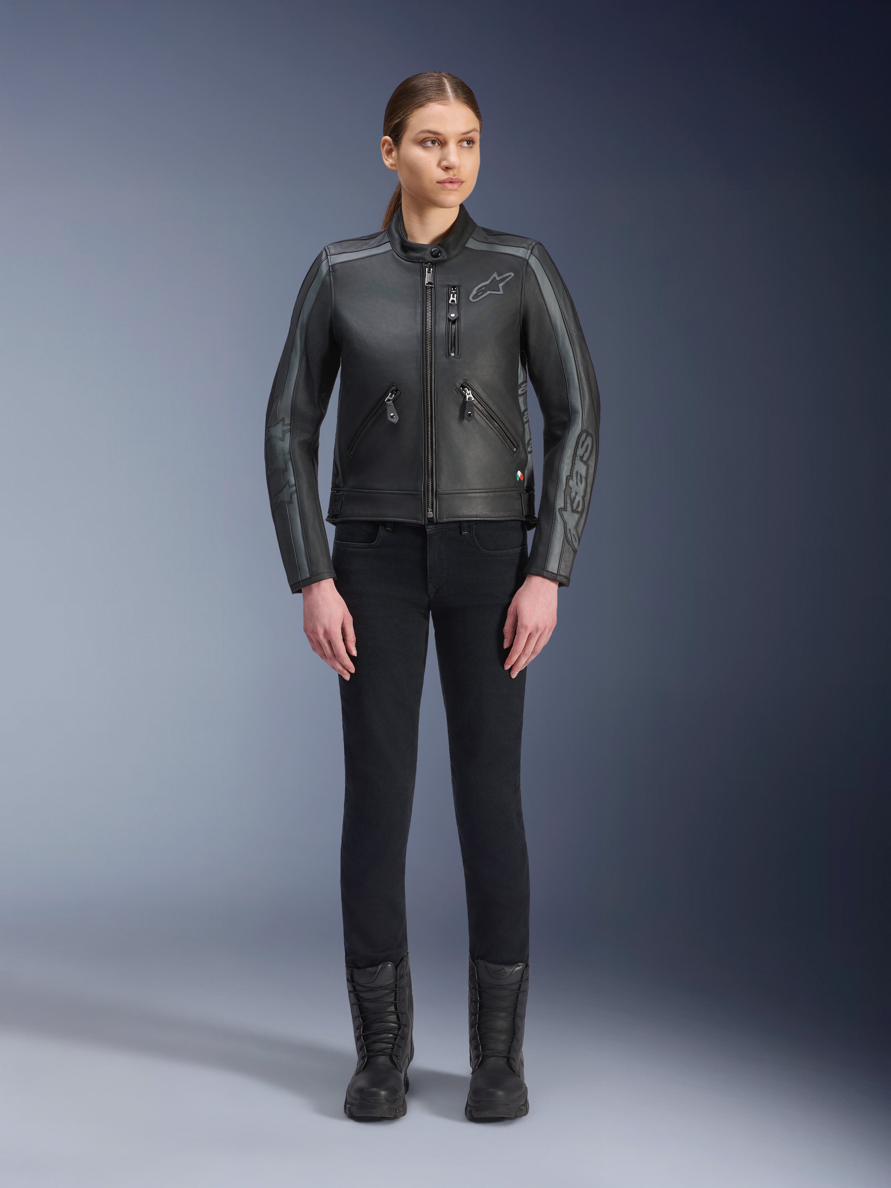 Alpinestars Stella Dyno Leather Jacket, damska kurtka motocyklowa, czarna z szarymi i ciemnoszarymi akcentami, studyjny widok całej sylwetki modelki w czarnych spodniach typu slim-fit i czarnych butach, wykonana ze skóry bydlęcej z subtelnym logo na piersi i rękawach