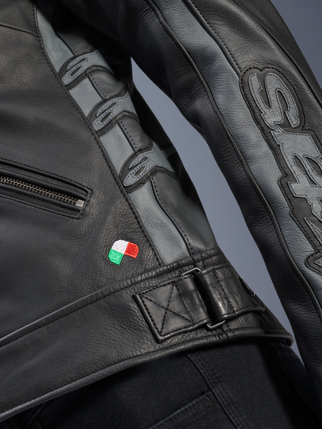 Alpinestars Stella Dyno Leather Jacket, detal, czarna i szara (Tar Gray) skóra bydlęca, zbliżenie na mały haft z flagą Włoch, regulowana klamra w pasie, szare skórzane panele z wytłoczonym logo Alpinestars i napisem Stella na rękawie.
